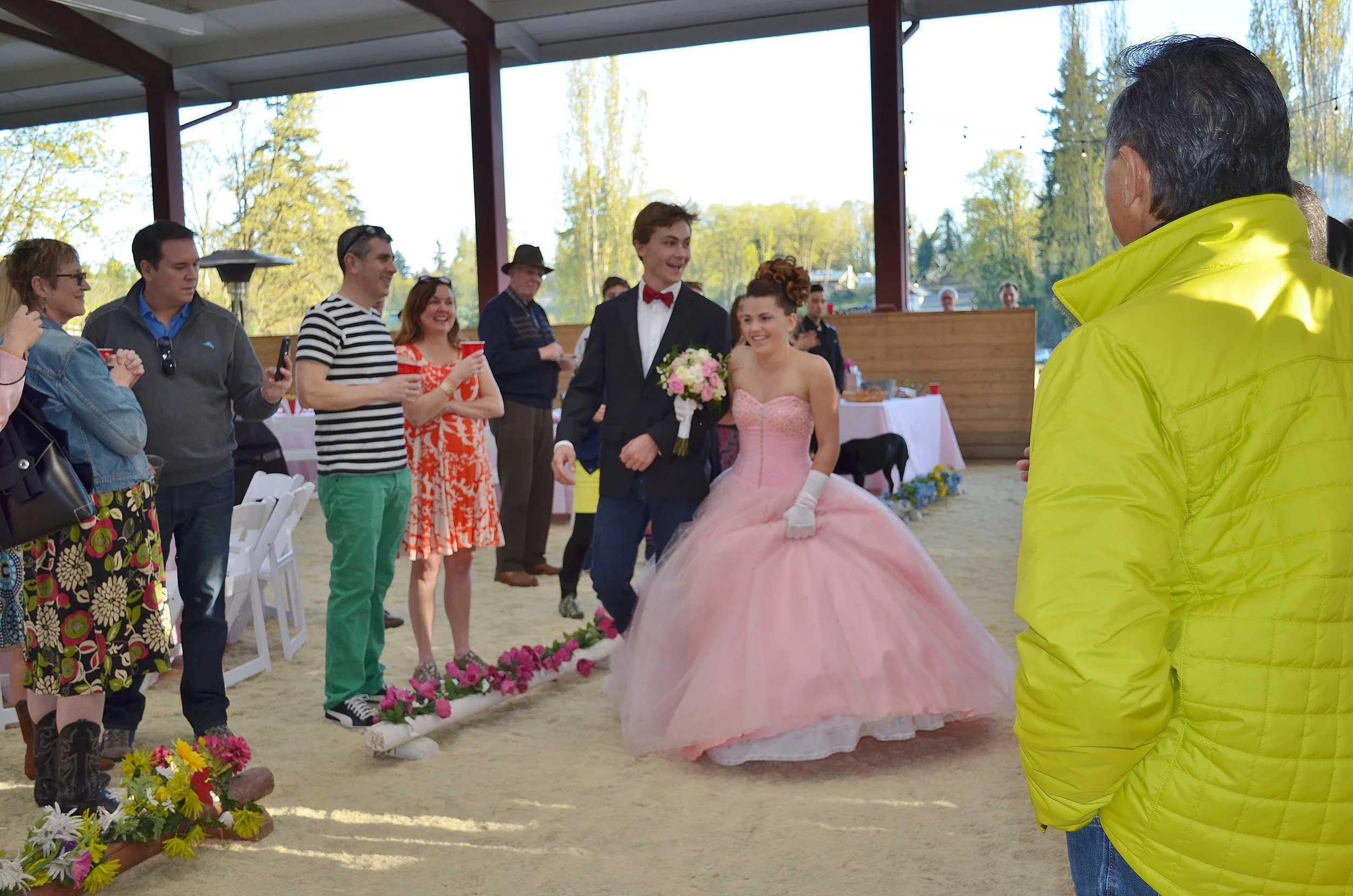 A couple dressed in wedding attire, with the groom in a tuxedo and the bride in a pink gown, walking arm in arm and smiling at each other at a wedding reception, surrounded by seated and standing guests.