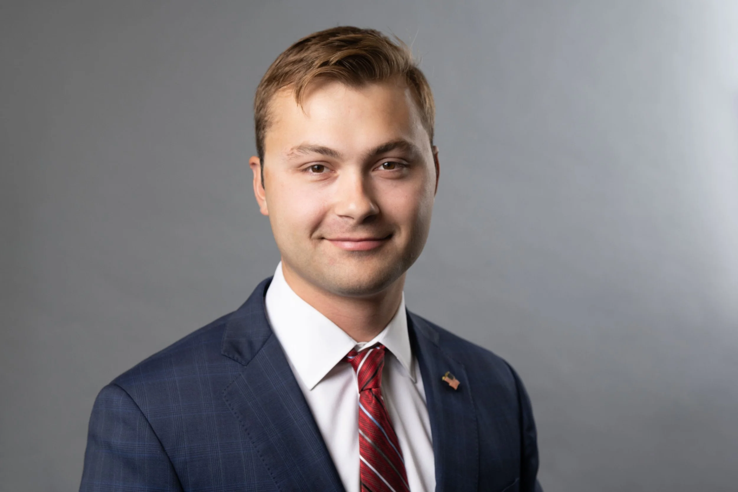 Young man in a navy suit with a red striped tie, wearing a small American flag lapel pin, smiling against a neutral gray background.