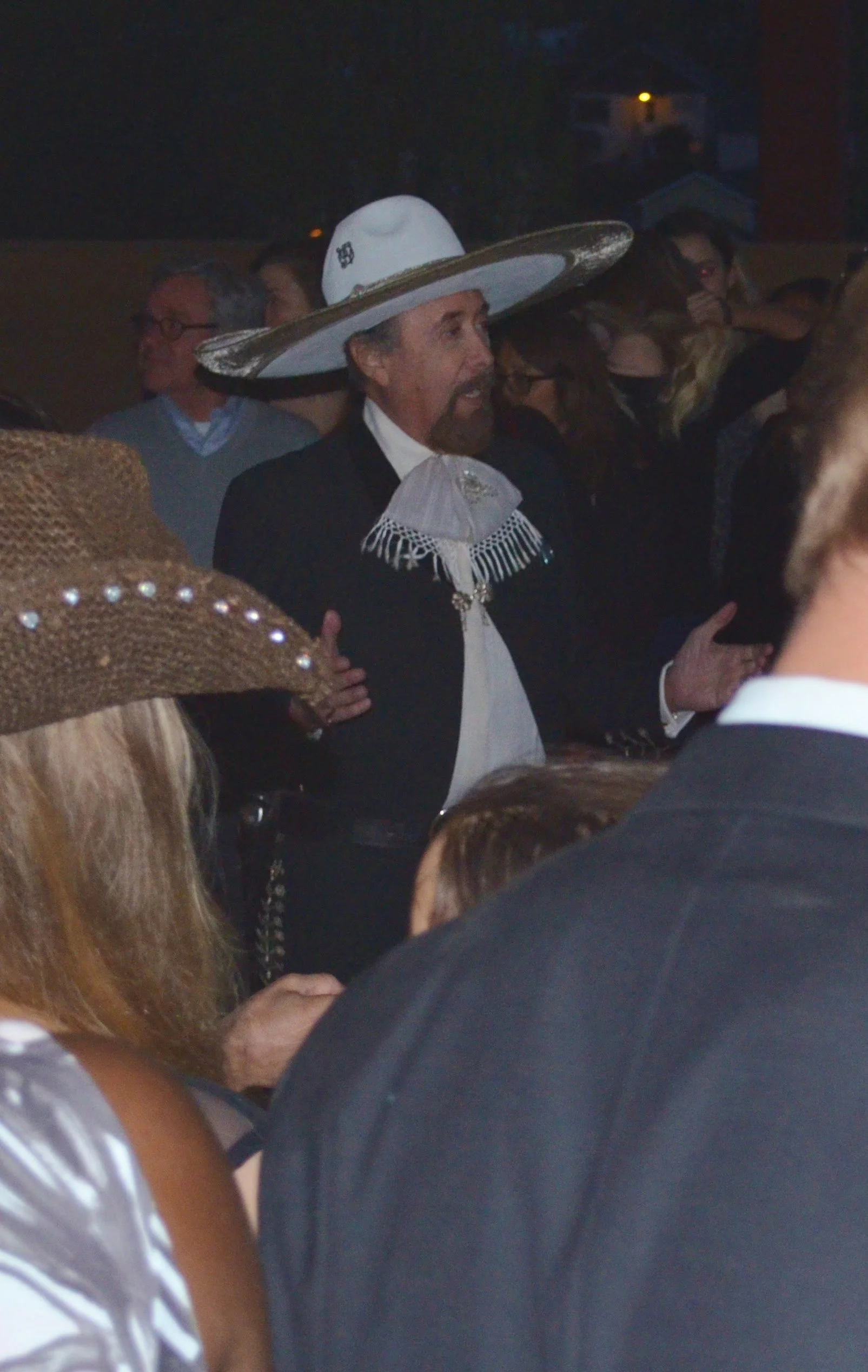 A man wearing a large white sombrero, a traditional Mexican charro suit, and a lacy necktie, appears to be dancing or celebrating at a crowded event.
