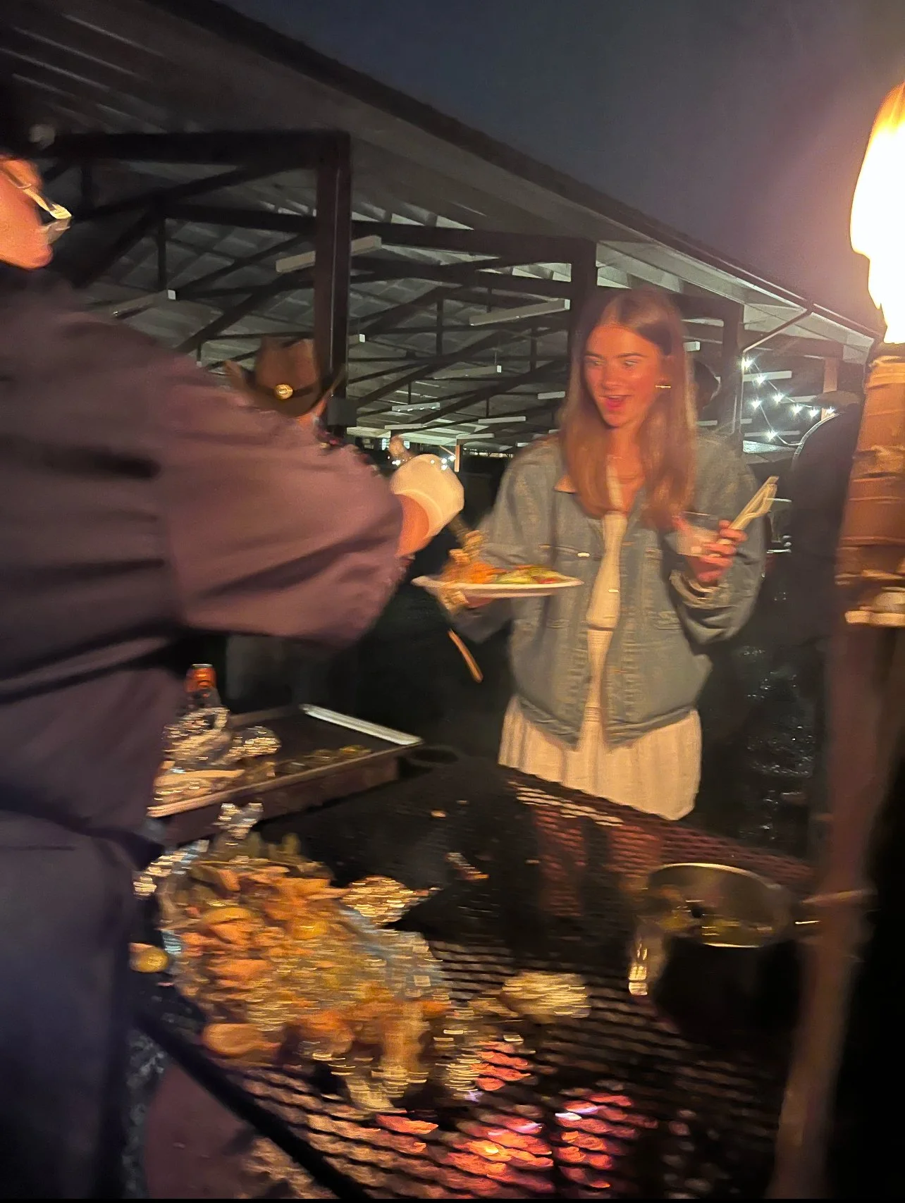 A young woman is smiling while holding a plate of food and chopsticks, standing near a grill with food being cooked, with a person serving her at an outdoor night gathering.