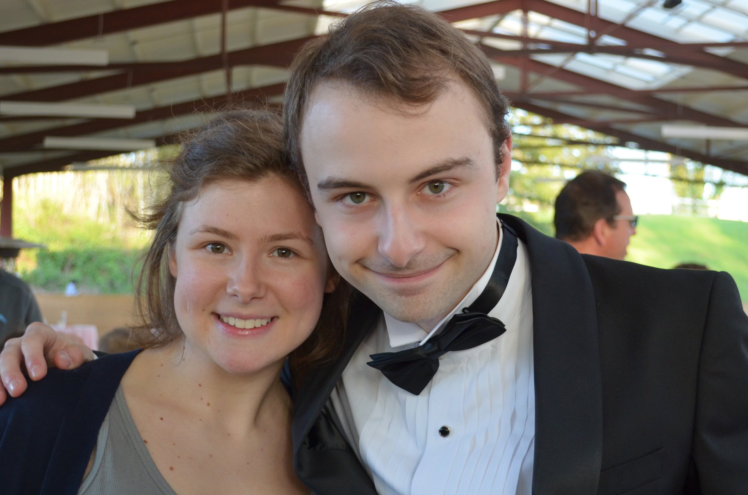 A young woman and a young man in formal attire smiling and posing closely together at an outdoor event under a canopy, with other people in the background.