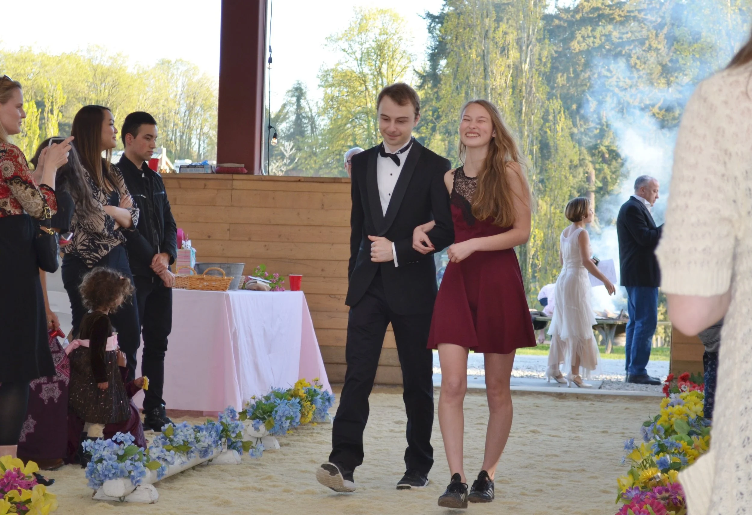 Young man in tuxedo and young woman in burgundy dress walking arm in arm at a formal event with guests watching. The background shows a wood-paneled wall, large windows with trees outside, and a table with pink tablecloth and flowers.