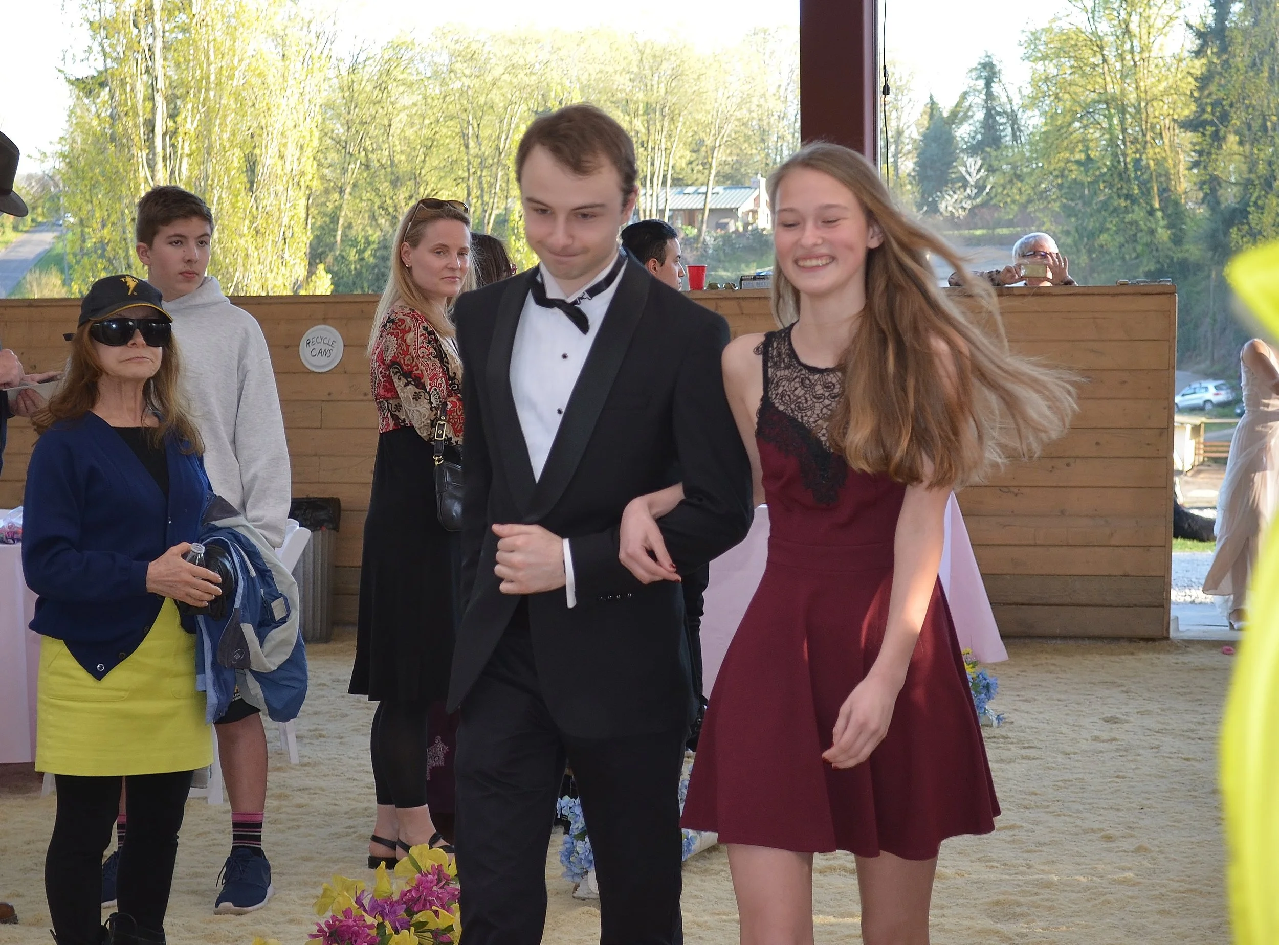 A young man in a black tuxedo and a young woman in a burgundy dress walking arm-in-arm, smiling, at a social event indoors with people in the background.