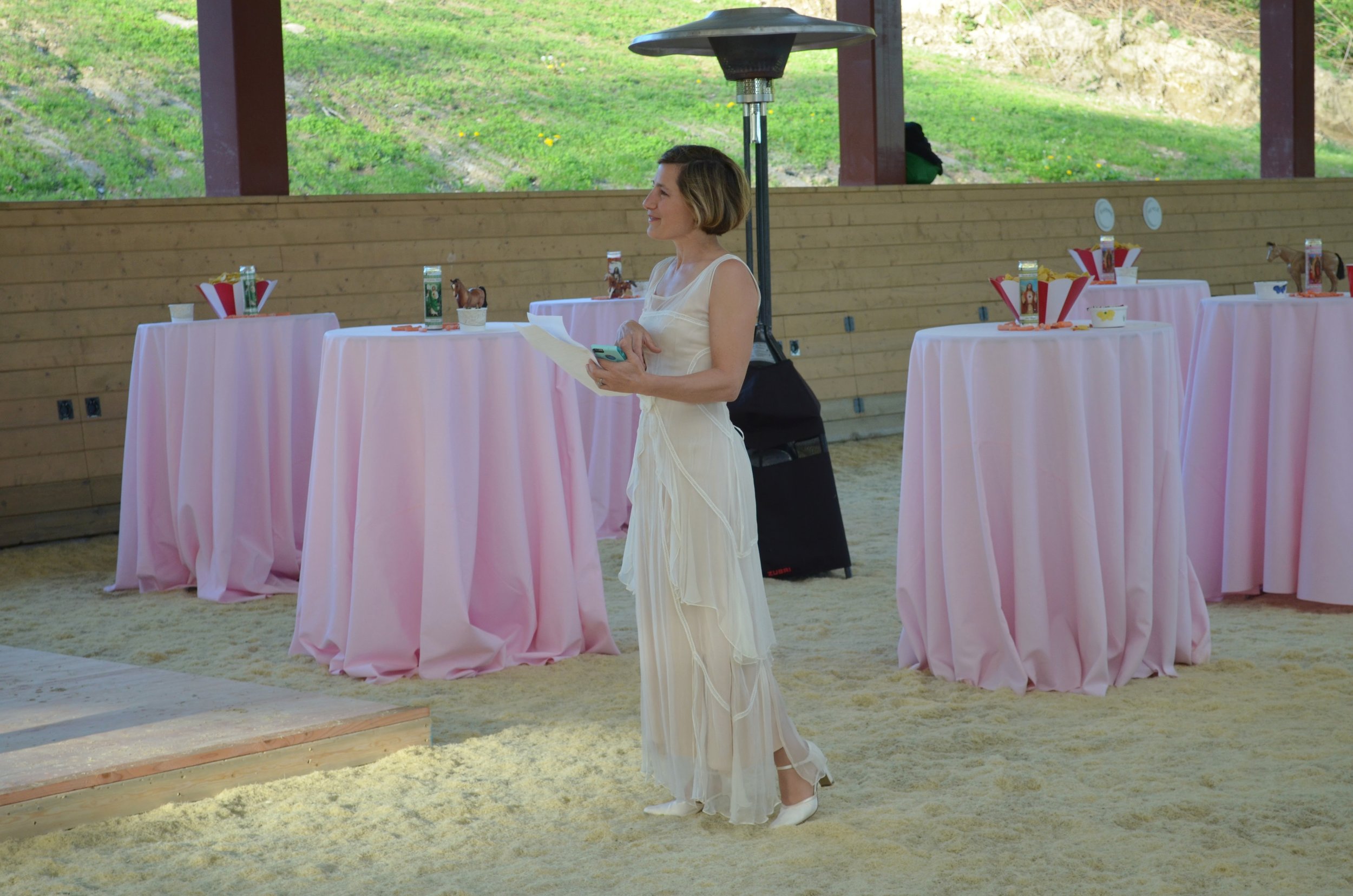 A woman in a white dress holding papers and a phone, standing in a decorated event space with pink tablecloths and presents on tables, with a wooden wall and large windows showing outdoors.