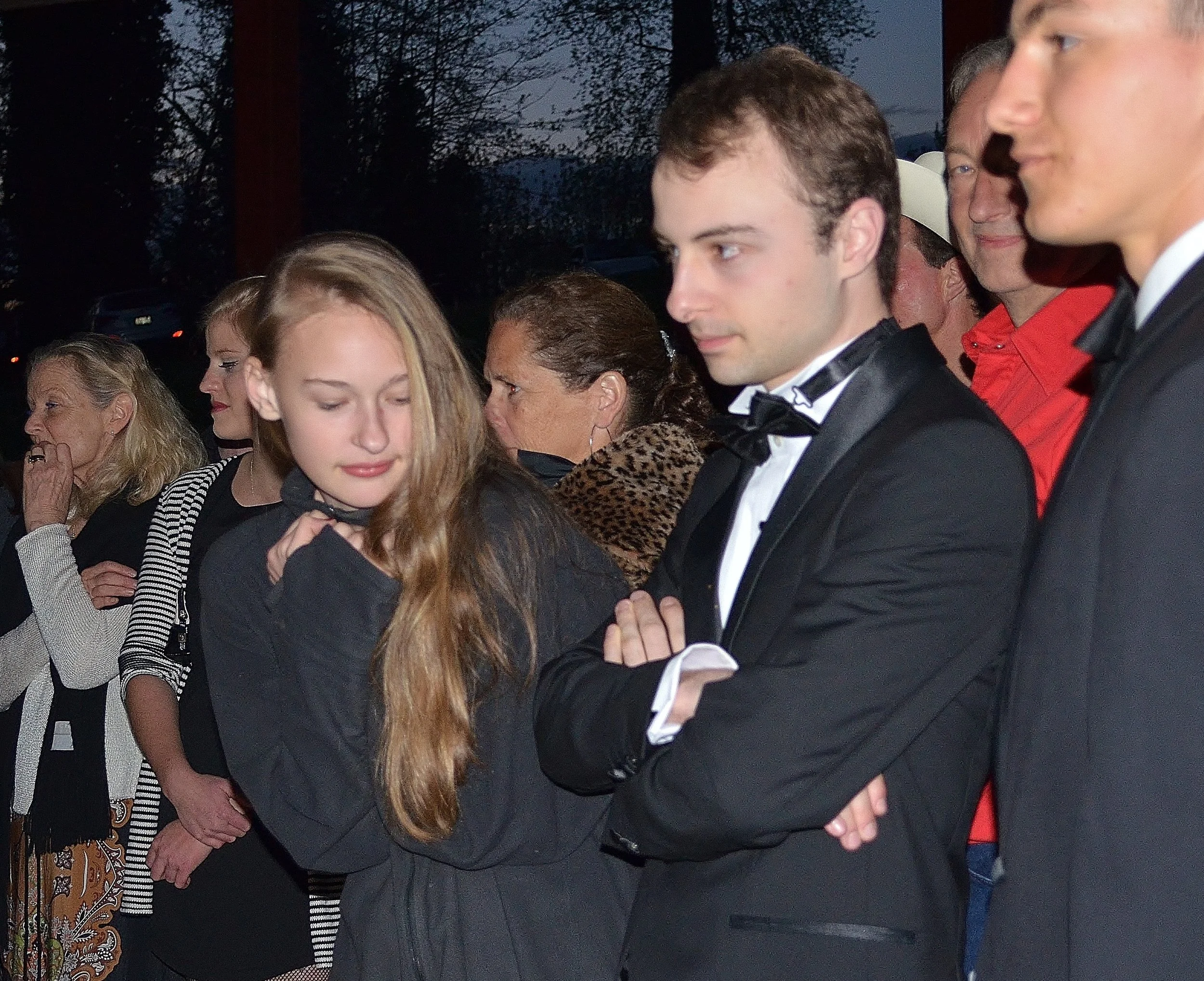 A group of people standing in a line at an outdoor event during evening, dressed in formal attire including tuxedos and dresses, some with serious or contemplative expressions.
