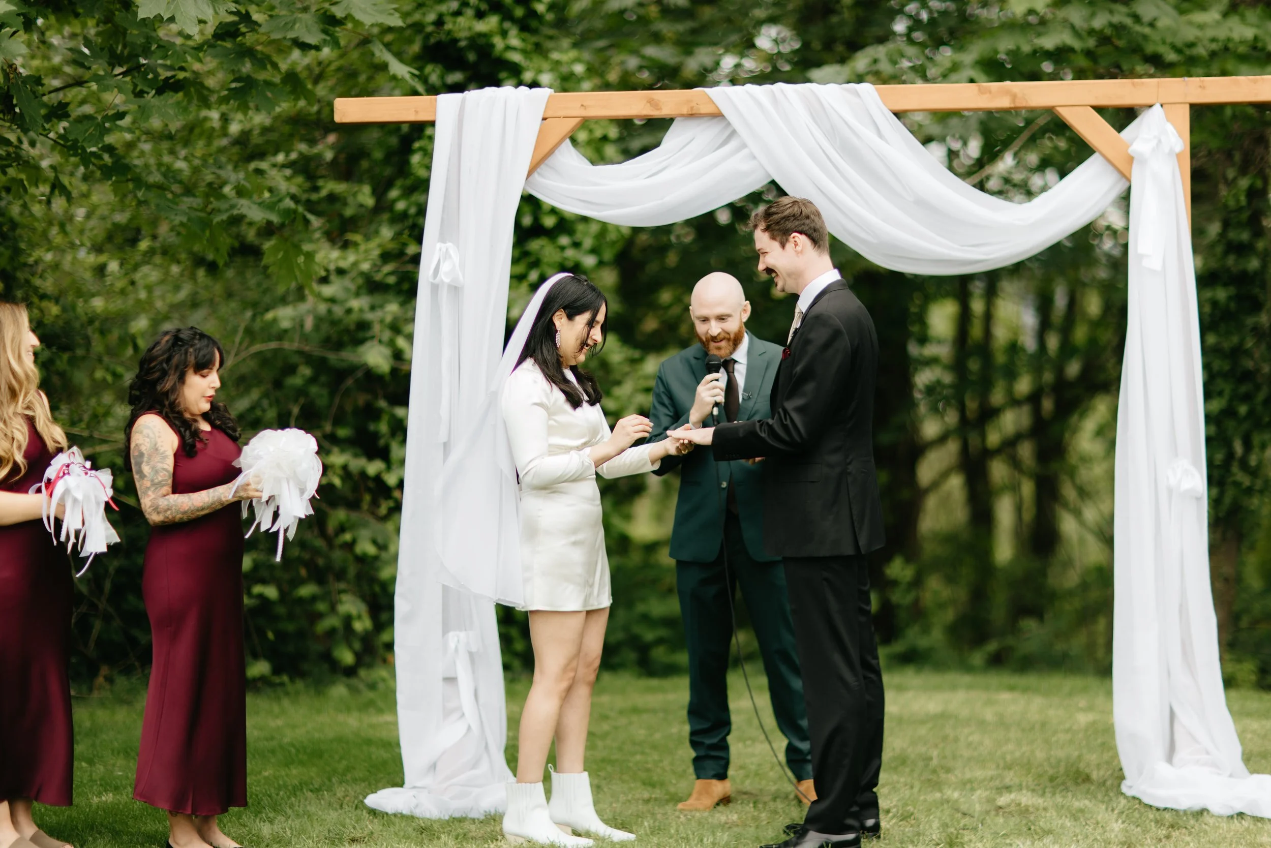 A couple exchanging wedding rings during an outdoor ceremony under a white fabric draped arch, with a bride and groom in formal attire, officiant, and bridesmaids holding bouquets, surrounded by green trees.