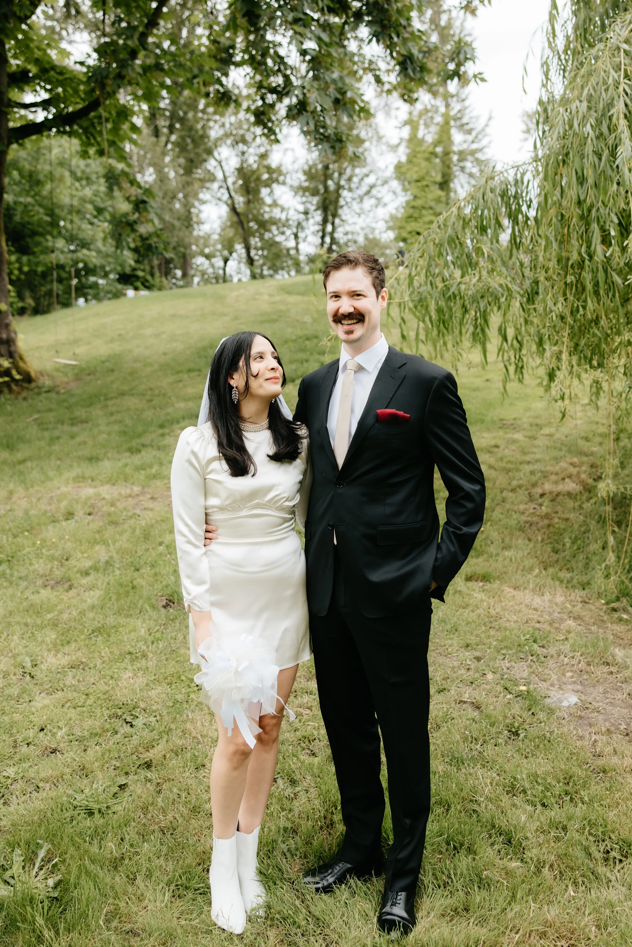 A couple dressed in wedding attire standing outdoors on grass, with trees in the background. The woman wears a white dress and white boots, and the man wears a black suit with a white shirt and red pocket square. They are smiling and standing close t