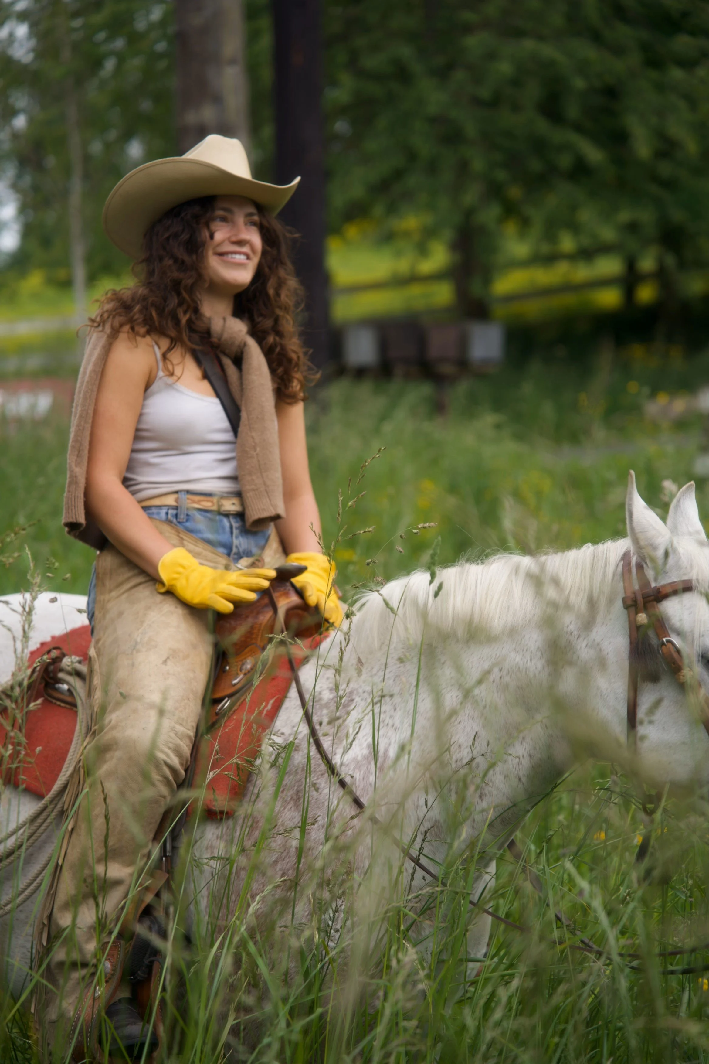 A woman wearing a cowboy hat, yellow gloves, and a beige scarf sits on a white horse in a grassy field with trees in the background.