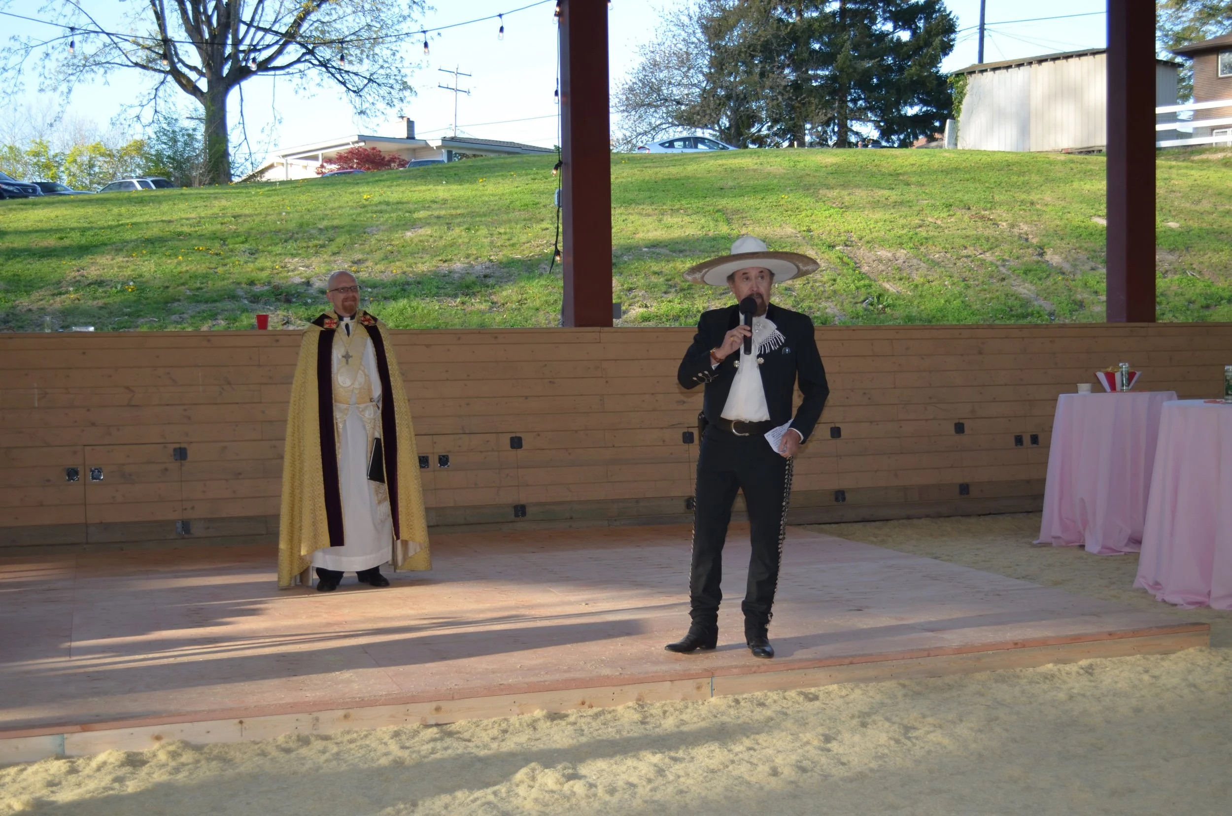 Two men in traditional Mexican costumes standing on a wooden stage outdoors. One man is dressed as a priest, and the other is wearing a charro suit with a large sombrero, speaking into a microphone. There are pink tablecloths on tables to the right a