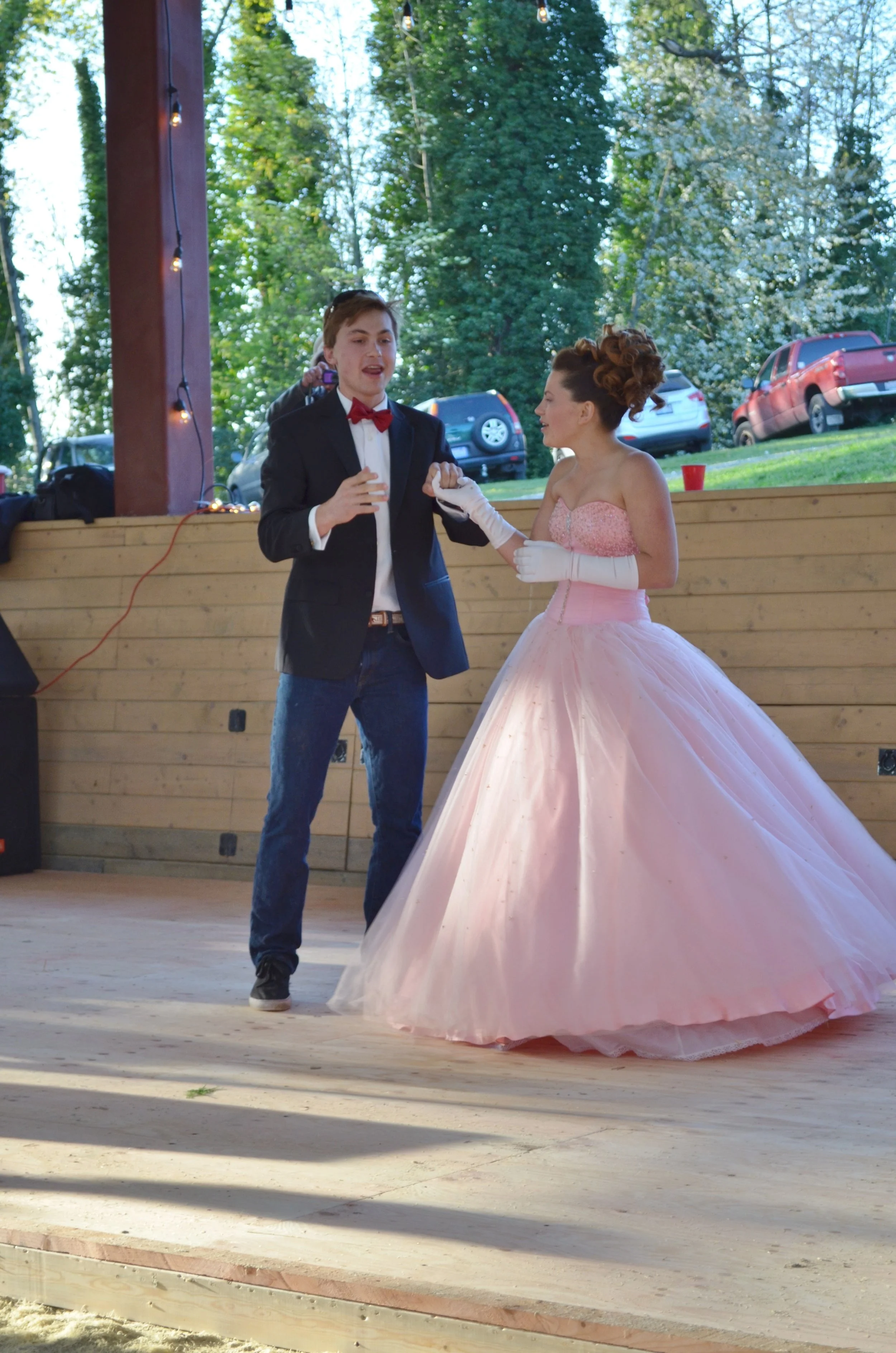 A young man in a tuxedo and a young woman in a pink ball gown are dancing together at an outdoor event. The woman is wearing white gloves and has curly hair. There are cars parked in the background, and string lights are hanging from a wooden structu