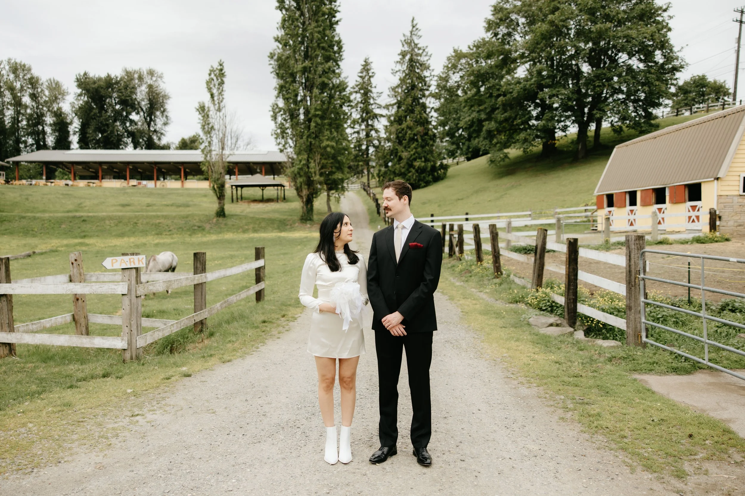 A couple dressed in vintage-style wedding attire standing on a gravel path in a rural farm setting, with a fence, grassy fields, trees, a horse, and farm buildings in the background.