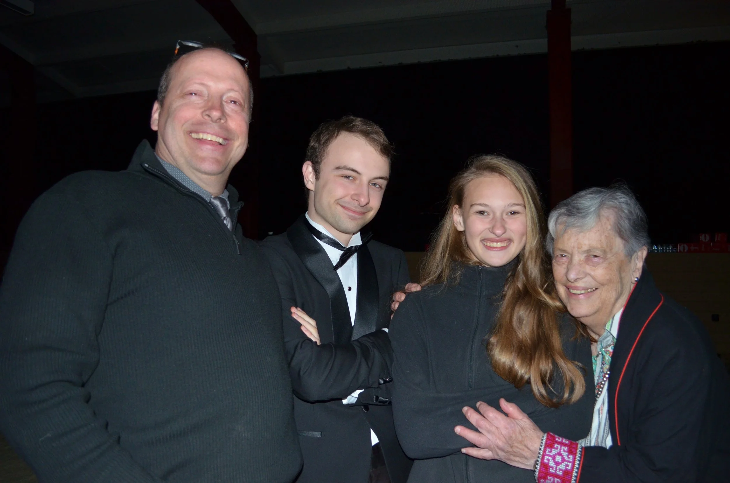 Four people smiling together at a social event, with one man in a black sweater, a young man in a tuxedo, a young woman in a black jacket, and an elderly woman in a black blazer with a colorful scarf, standing close and facing the camera.