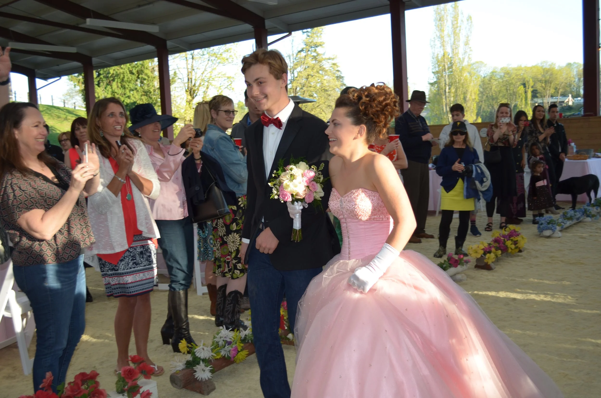 A young couple dressed as a bride and groom walking together, surrounded by smiling wedding guests in an outdoor setting with trees and sunlight.