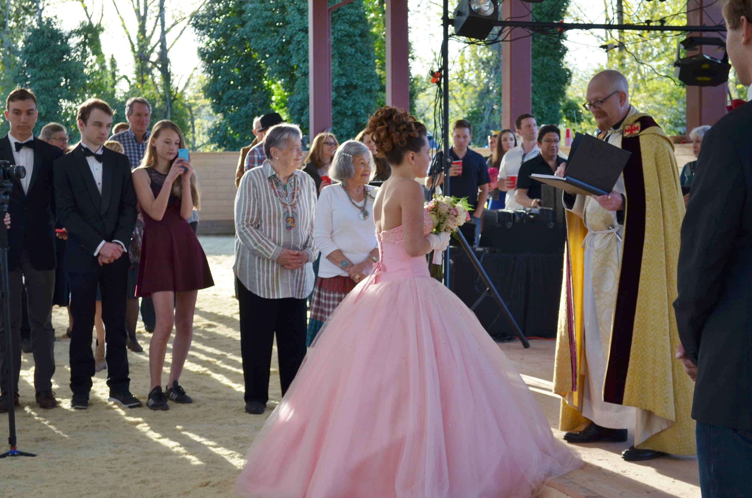 A young woman in a pink ball gown and gloves is participating in a ceremony, likely a coming-of-age or quinceañera, with an officiant in gold robes reading from a book. Several elders and young people in formal attire watch the event, with some takin