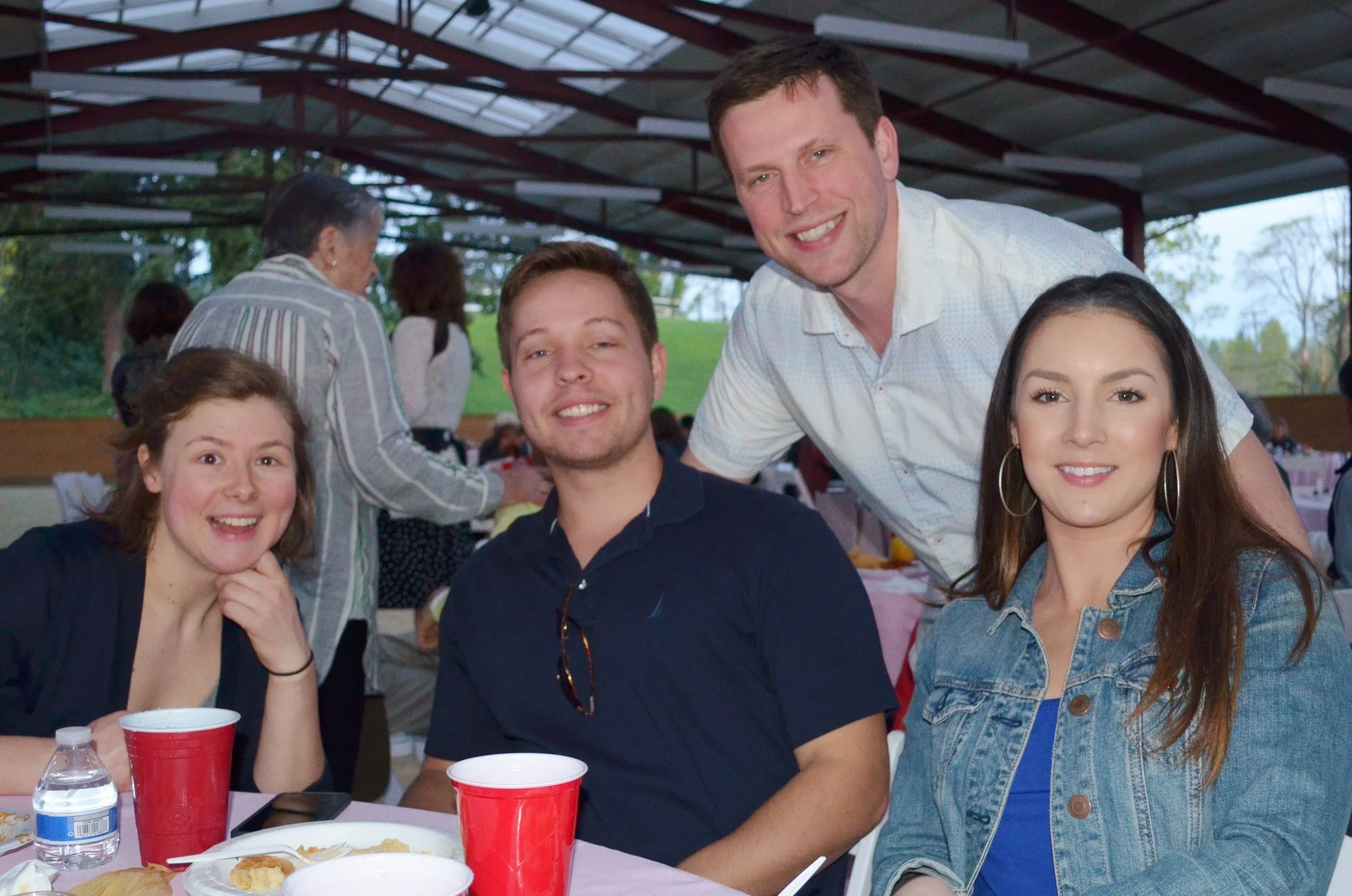 Group of four young adults at a social gathering, sitting at a table with red cups and food, smiling and looking at the camera, with a pavilion and people in the background.