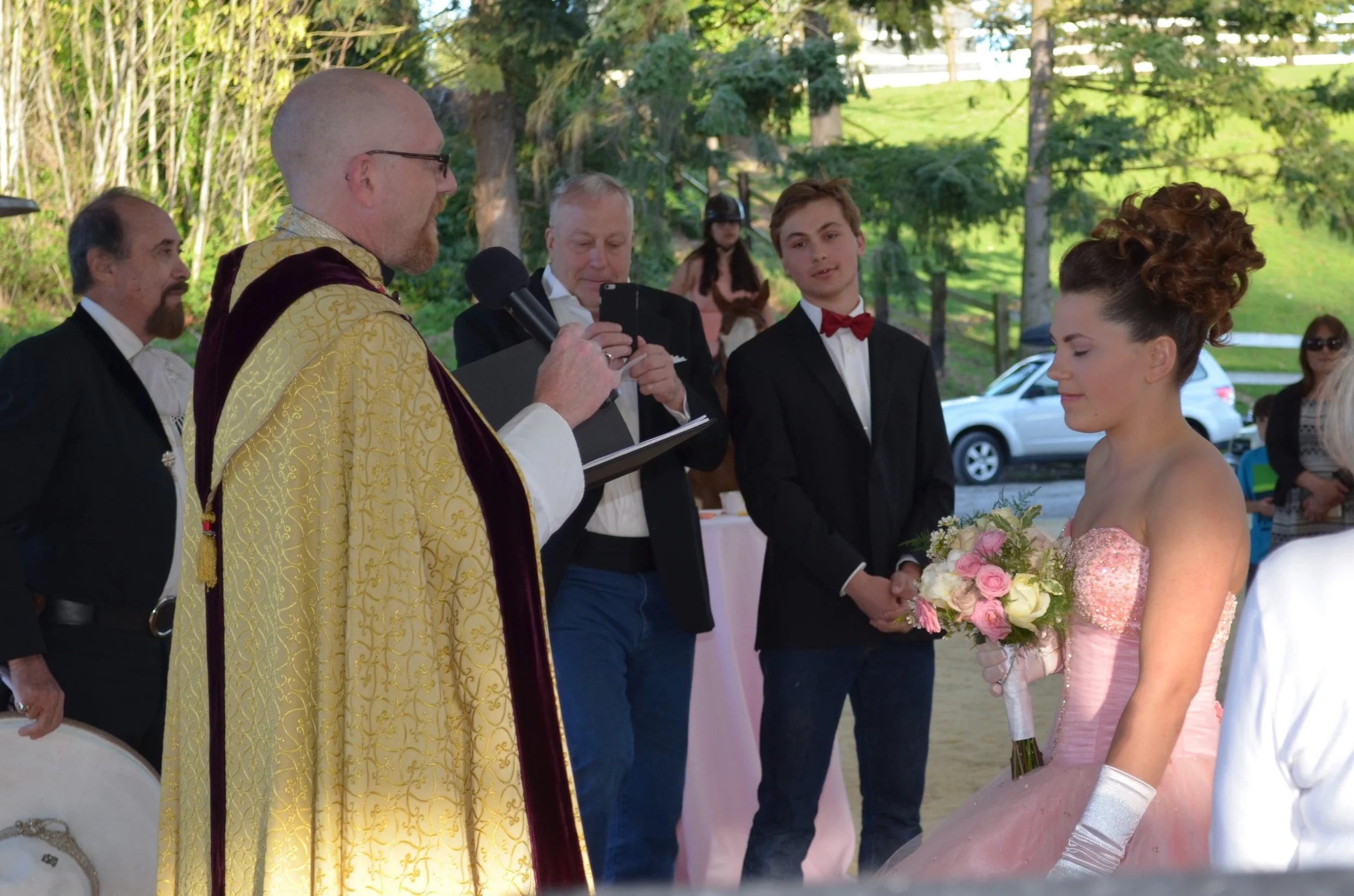 A wedding ceremony outdoors with a bride in a pink gown holding a bouquet of flowers and a groom in a tuxedo, surrounded by guests and an officiant