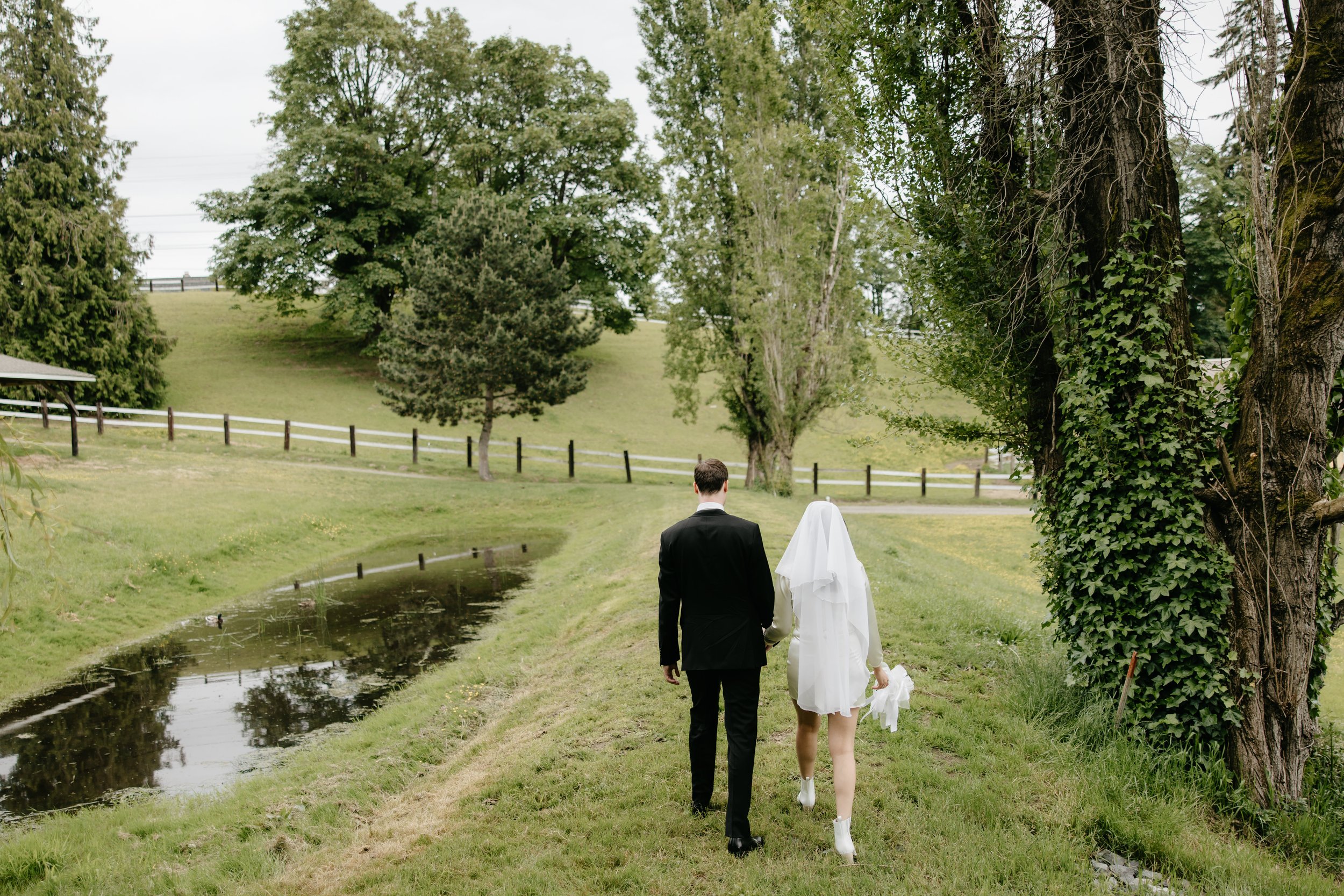 A couple dressed in wedding attire walking along a grassy path near a small canal, with trees and a hill in the background.