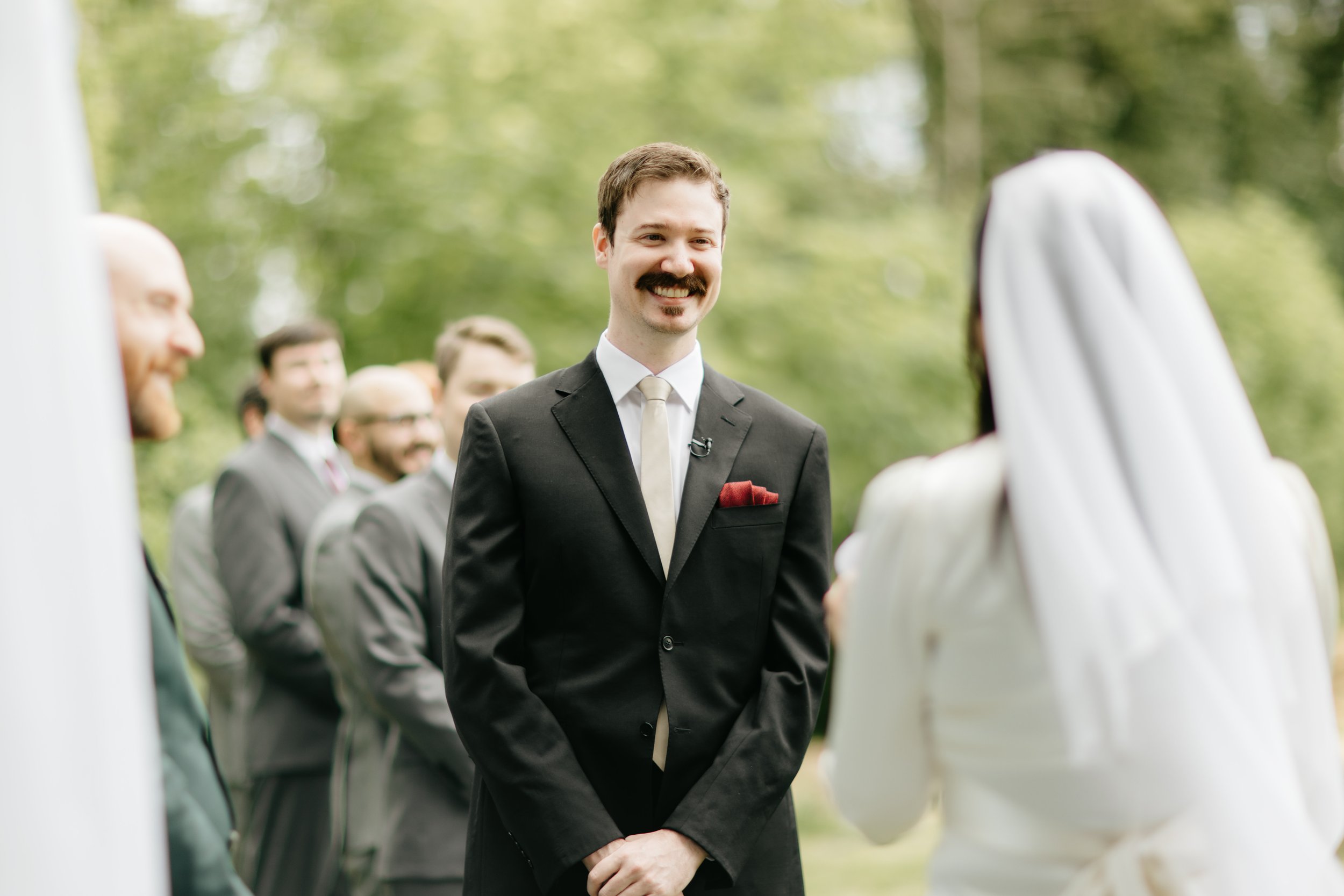 A groom with a mustache and dark suit standing outdoors during a wedding ceremony, smiling at the bride, with groomsmen in suits behind him and a bride in a white dress and veil in front of him.