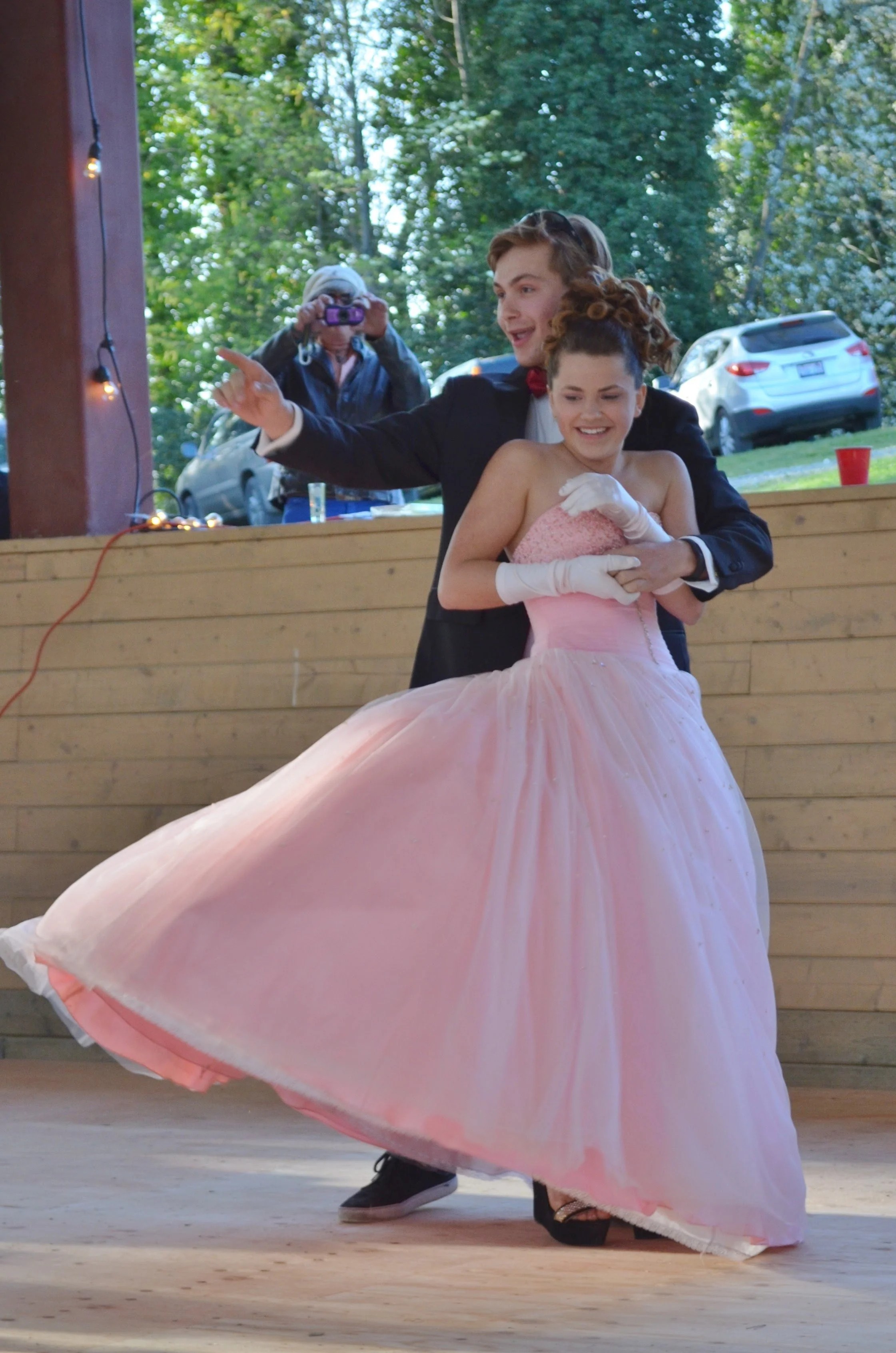 A young woman in a flowing pink gown being twirled around by a young man in a tuxedo, during a dance at an outdoor event. They are smiling and enjoying the moment, with an outdoor setting in the background.
