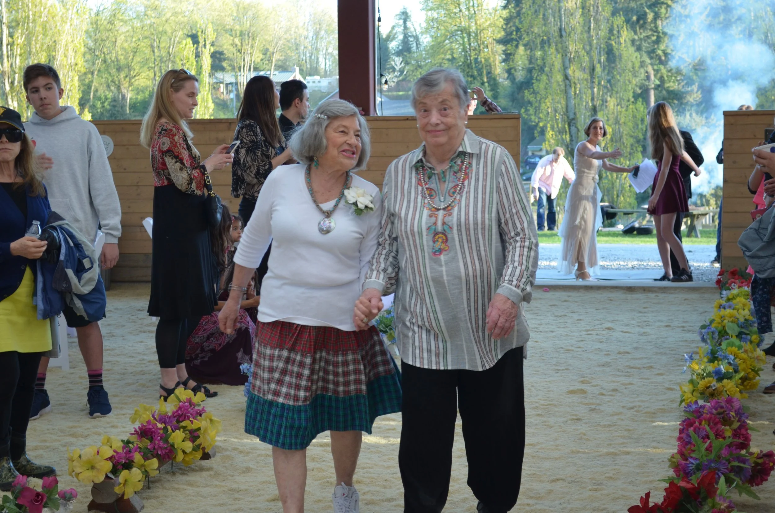 An elderly couple holds hands and walks through a decorated outdoor gathering, surrounded by various people, with trees and a blue sky in the background.