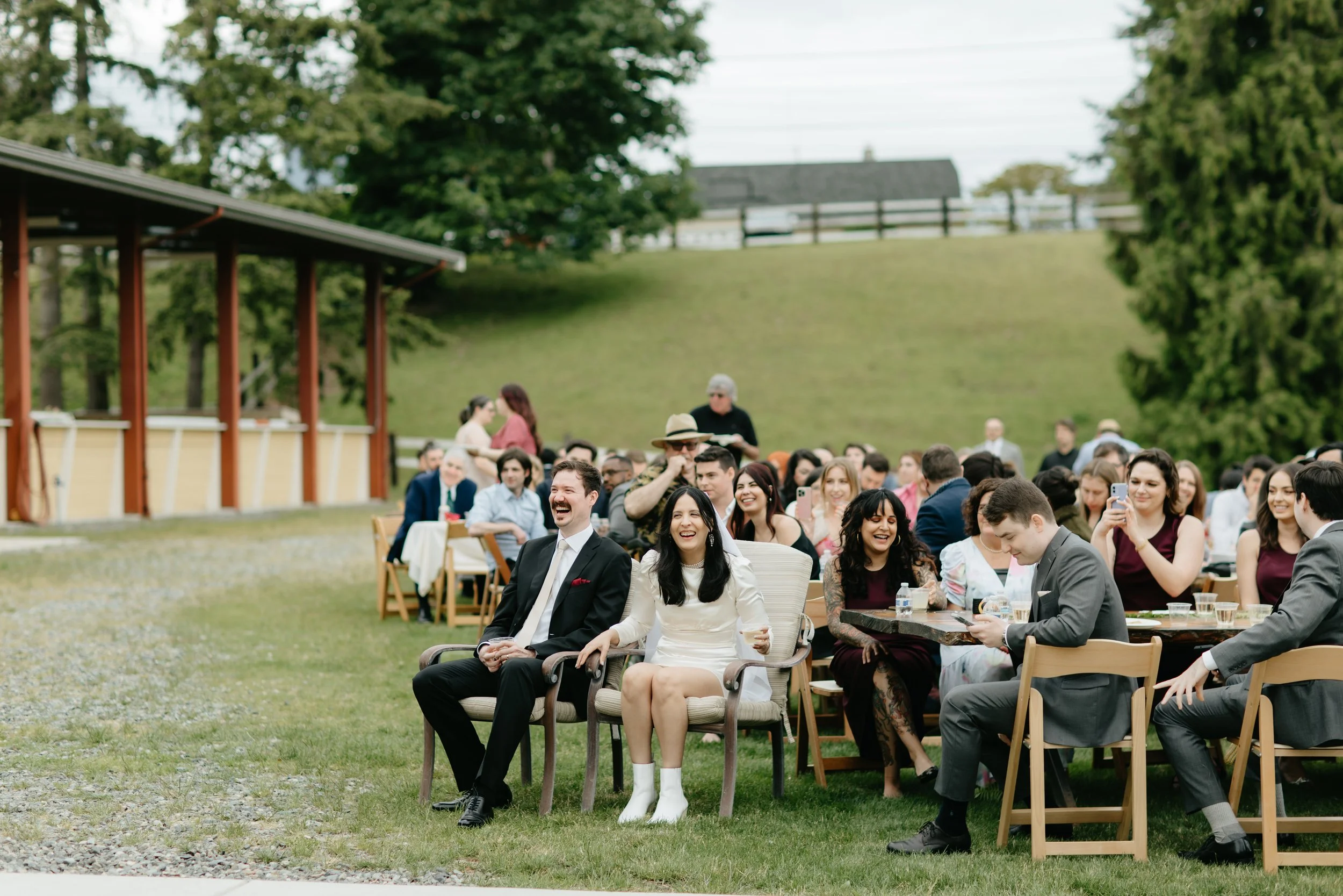 Guests seated outdoors at a wedding, some smiling and enjoying drinks, with a grassy hill and trees in the background.