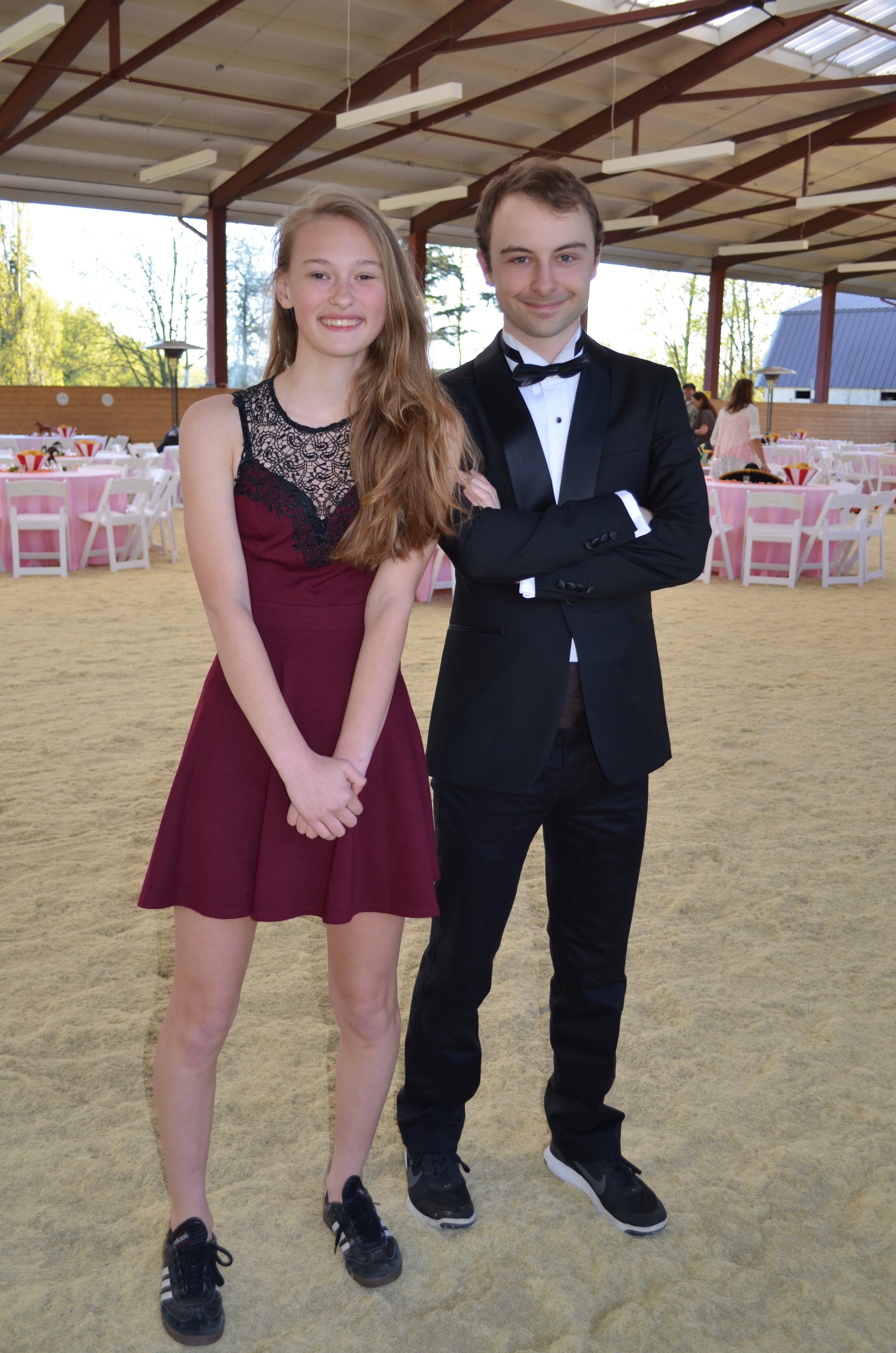 A young woman and man dressed formally at an indoor event with pink and white decorated tables in the background.