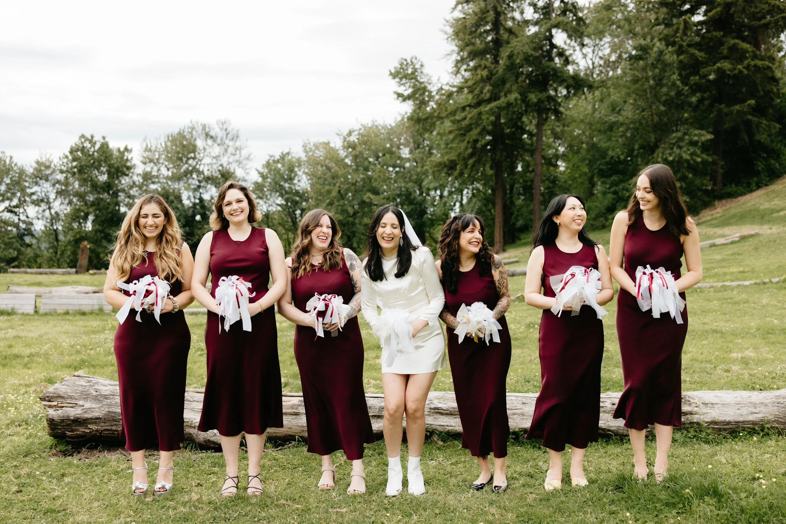 A group of seven women dressed for a wedding, standing outdoors on grass with trees in the background. The woman in the center is wearing a white dress and veil, while the others wear matching burgundy dresses. They are holding small bouquets or gift
