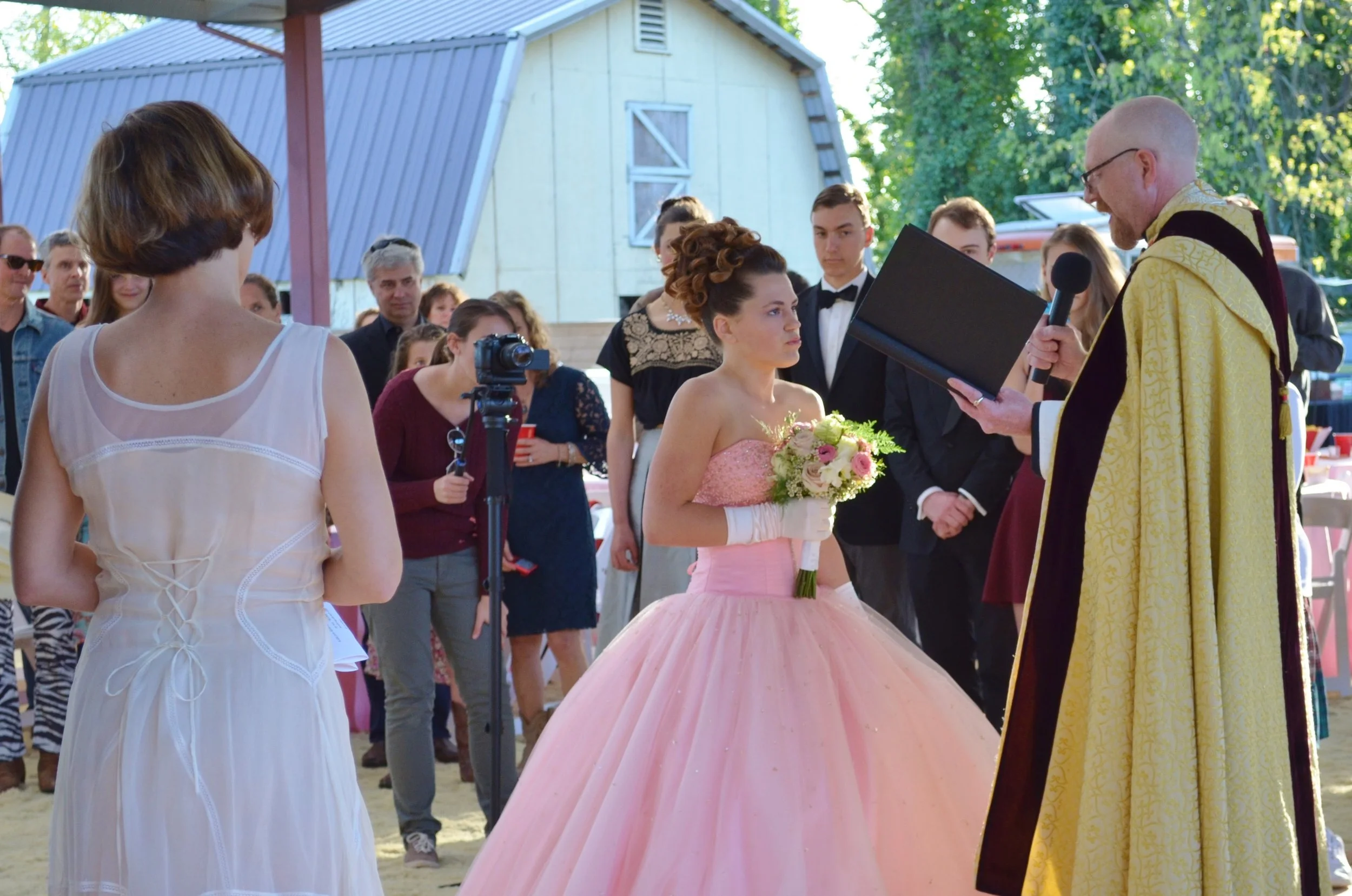 A young woman in a pink ball gown holding a bouquet and wearing white gloves standing before a man in religious robes reading from a book during a wedding ceremony outdoors.