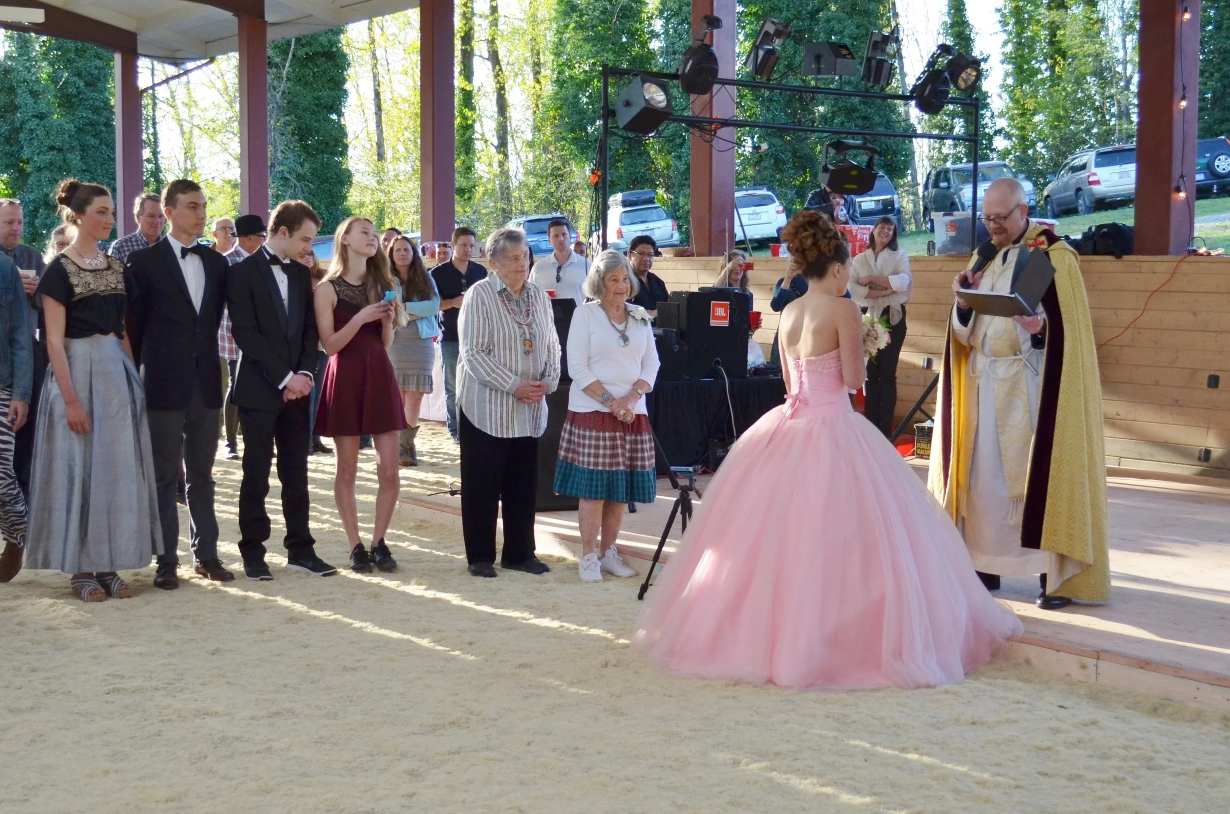 A wedding ceremony in an outdoor pavilion with a priest officiating, a bride in a pink wedding gown, and guests standing in line. The scene includes a wooded background and parking lot visible outside.