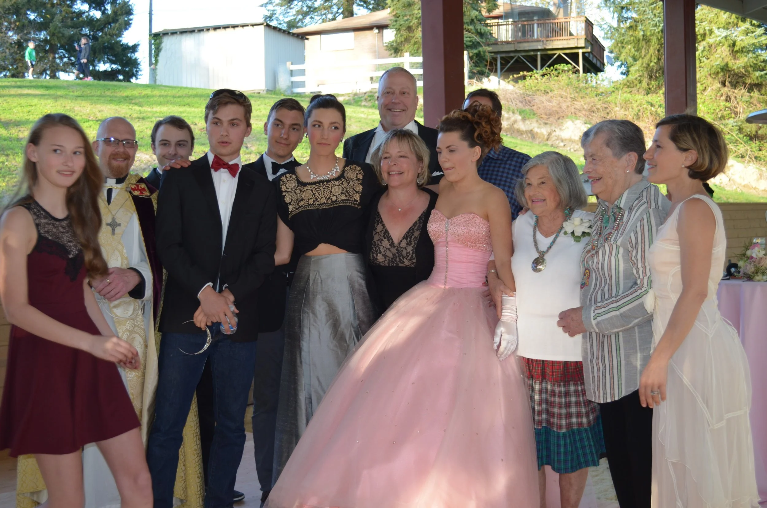 Group of people celebrating at a gathering, including teenagers in dresses and tuxedos, and elderly women in casual attire, standing outdoors under a pavilion with trees in the background.
