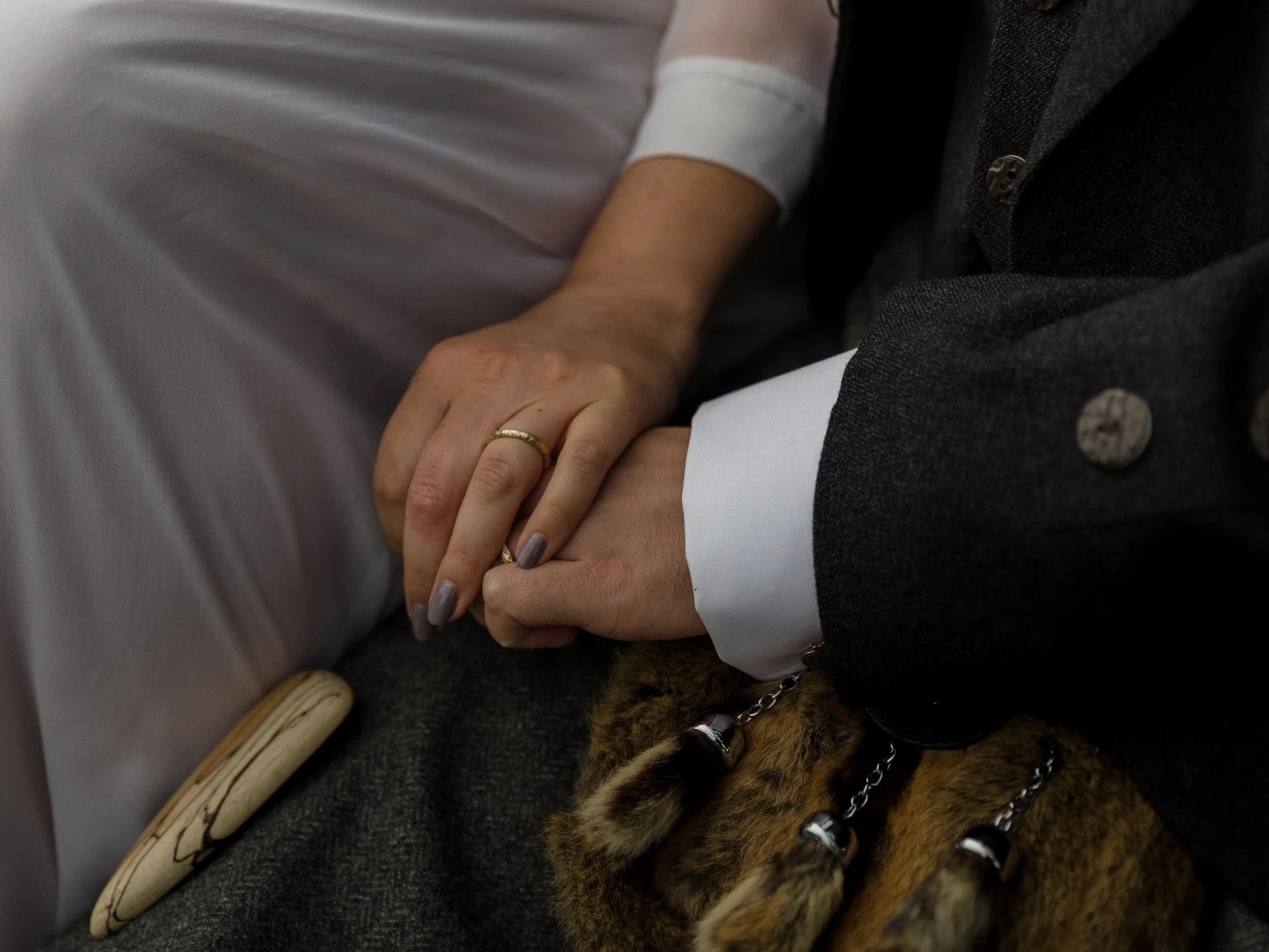 Close-up of a couple holding hands, with the woman wearing a gold wedding band and a white dress, and the man in traditional Scottish attire.