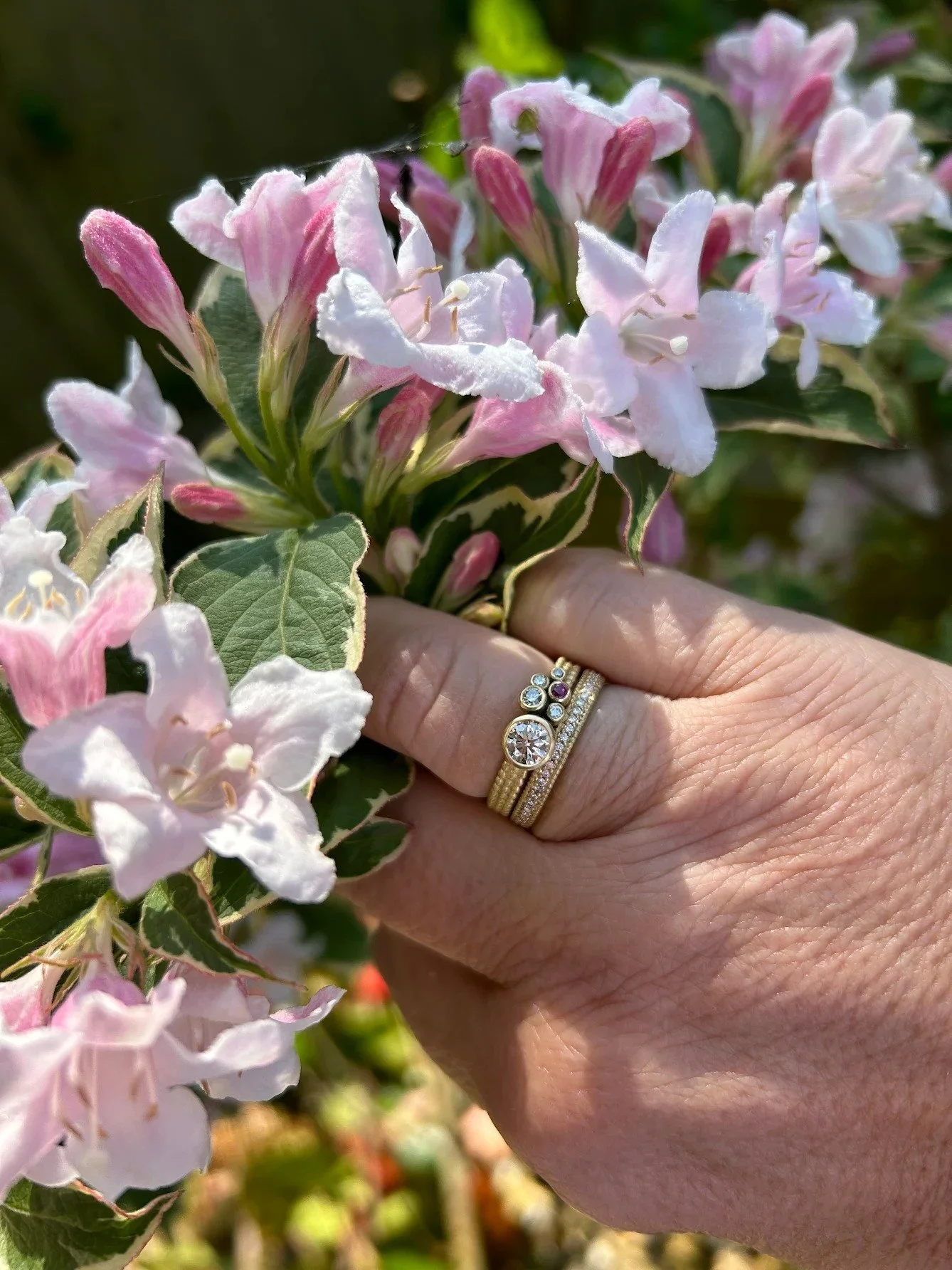 A hand adorned with a gold engagement ring featuring a large diamond, paired with delicate stacking rings, gently holding a bouquet of pink and white flowers.