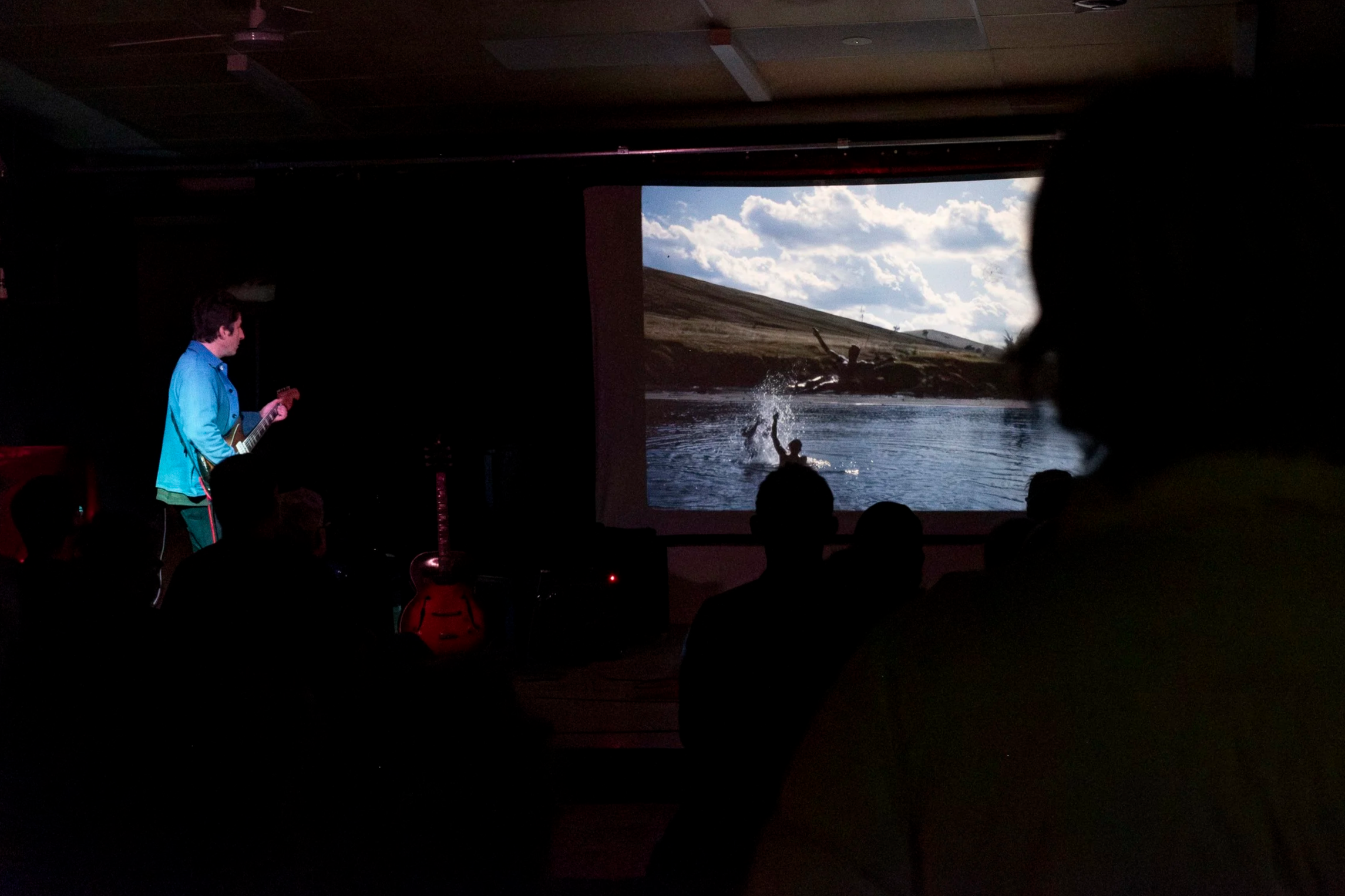 Spotlit guitarist in denim jacket onstage in a dark room, faces a large projection. Image on screen is of people skylarking in a dam.