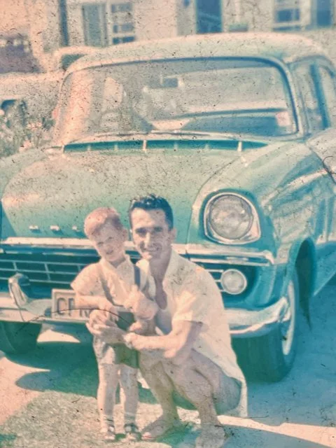 Smiling, dark-haired man and scowling red-haired child together in front of a 1960s Holden car. Image is a badly damaged slide.