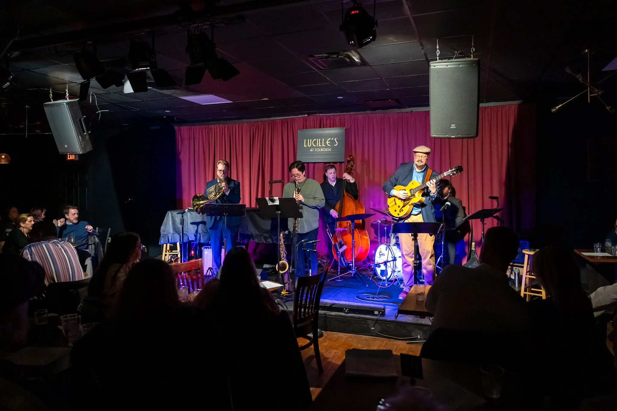 Quintet of musicians onstage playing french horn, soprano sax, upright bass, electric guitar, drums