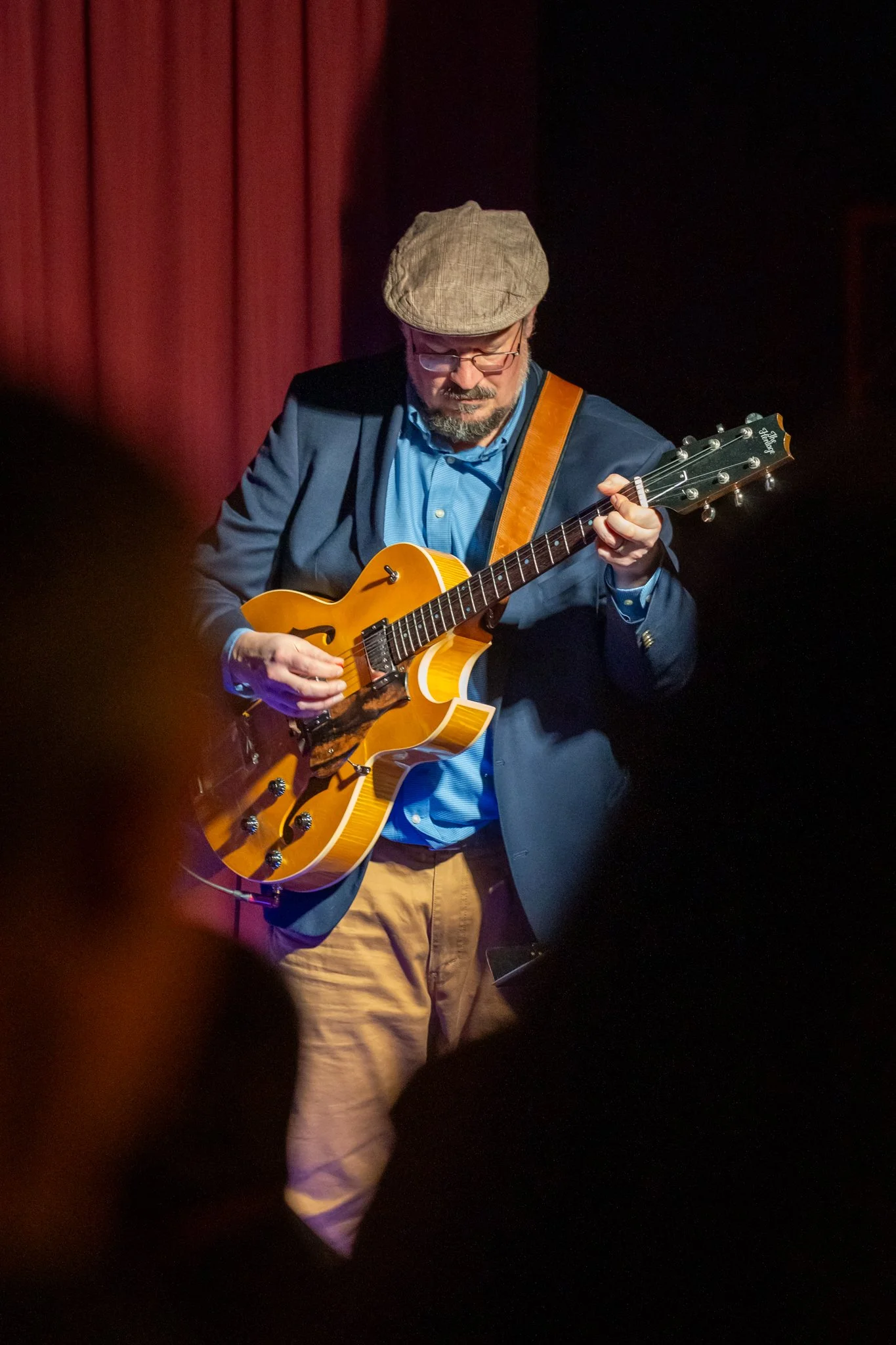 Man in hat with head down playing electric hollowbody guitar onstage.
