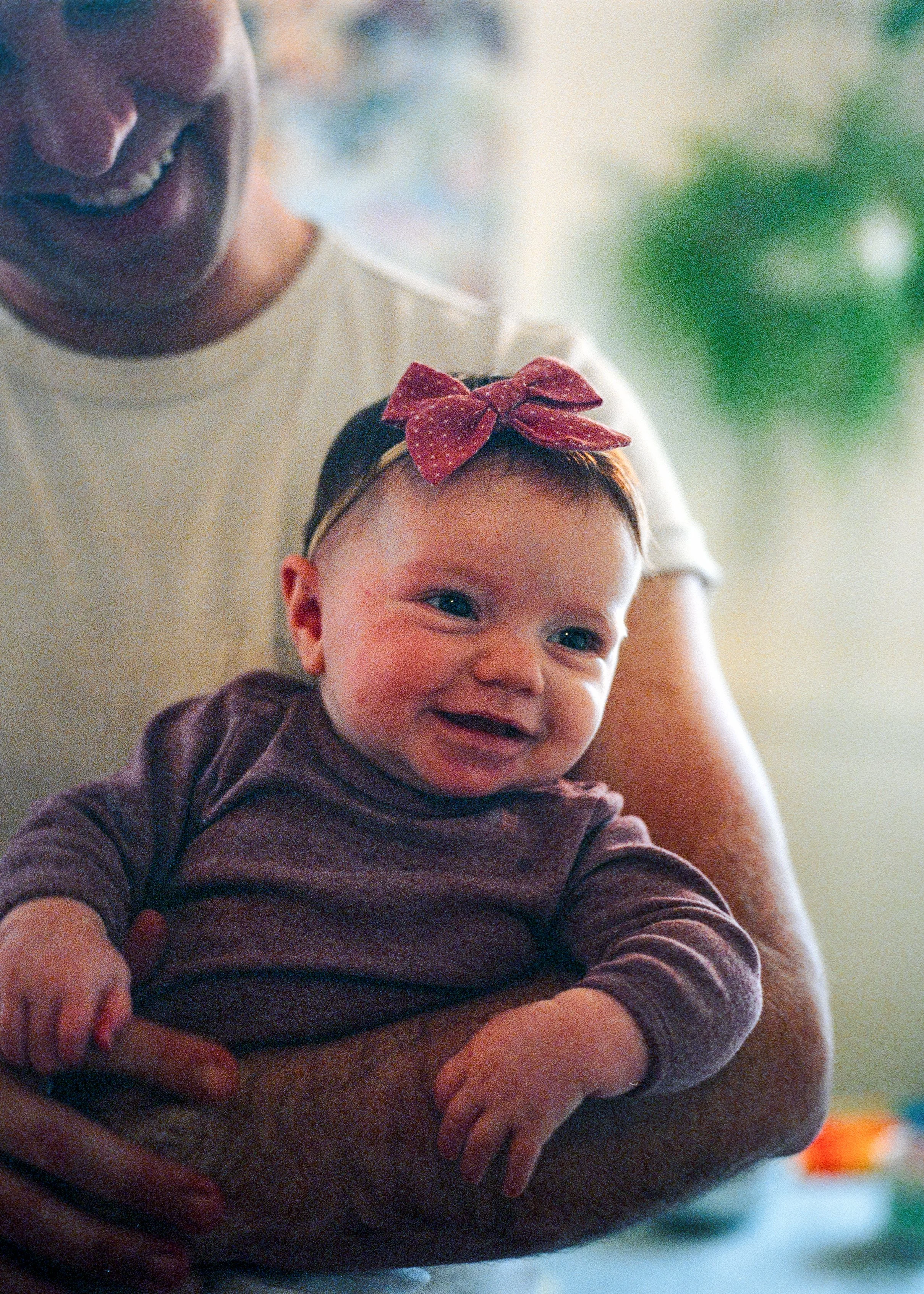 A documentary-style close-up portrait focusing on a happy baby with bright eyes and a large pink polka dot bow, being held securely by their father (face mostly out of frame). Warm colors and slight film grain, emphasizing natural family joy at home.