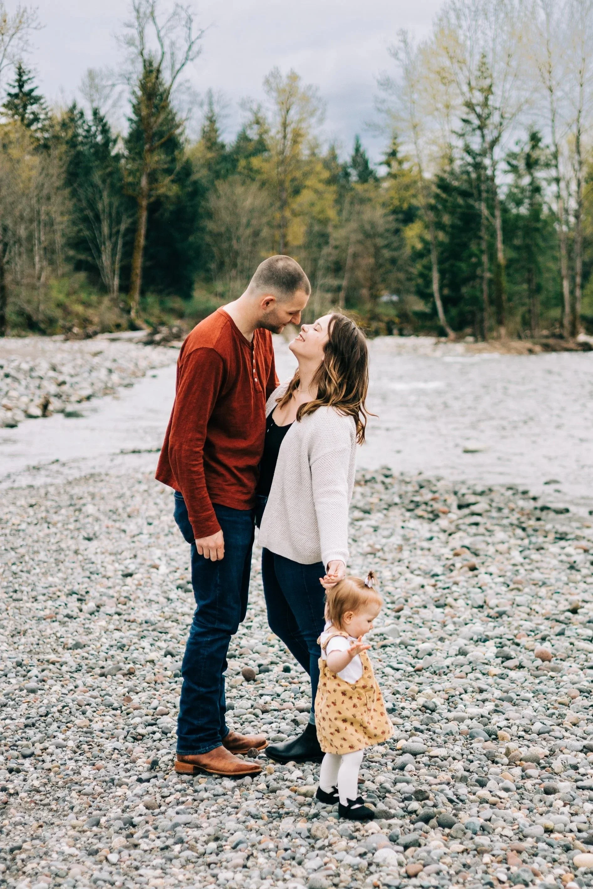 Mom and dad share a kiss by the river while holding their toddlers hand.