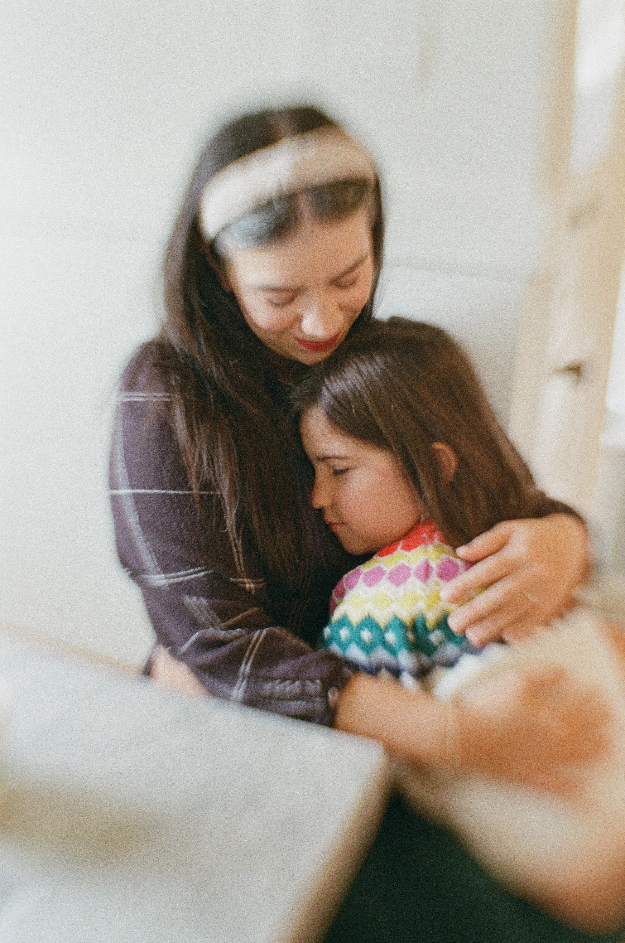 A loving mother with dark hair and a headband embraces her young daughter, whose face is buried in her mother’s chest, in a tender hug. Candid lifestyle family portrait photography with soft light and a gentle blur.