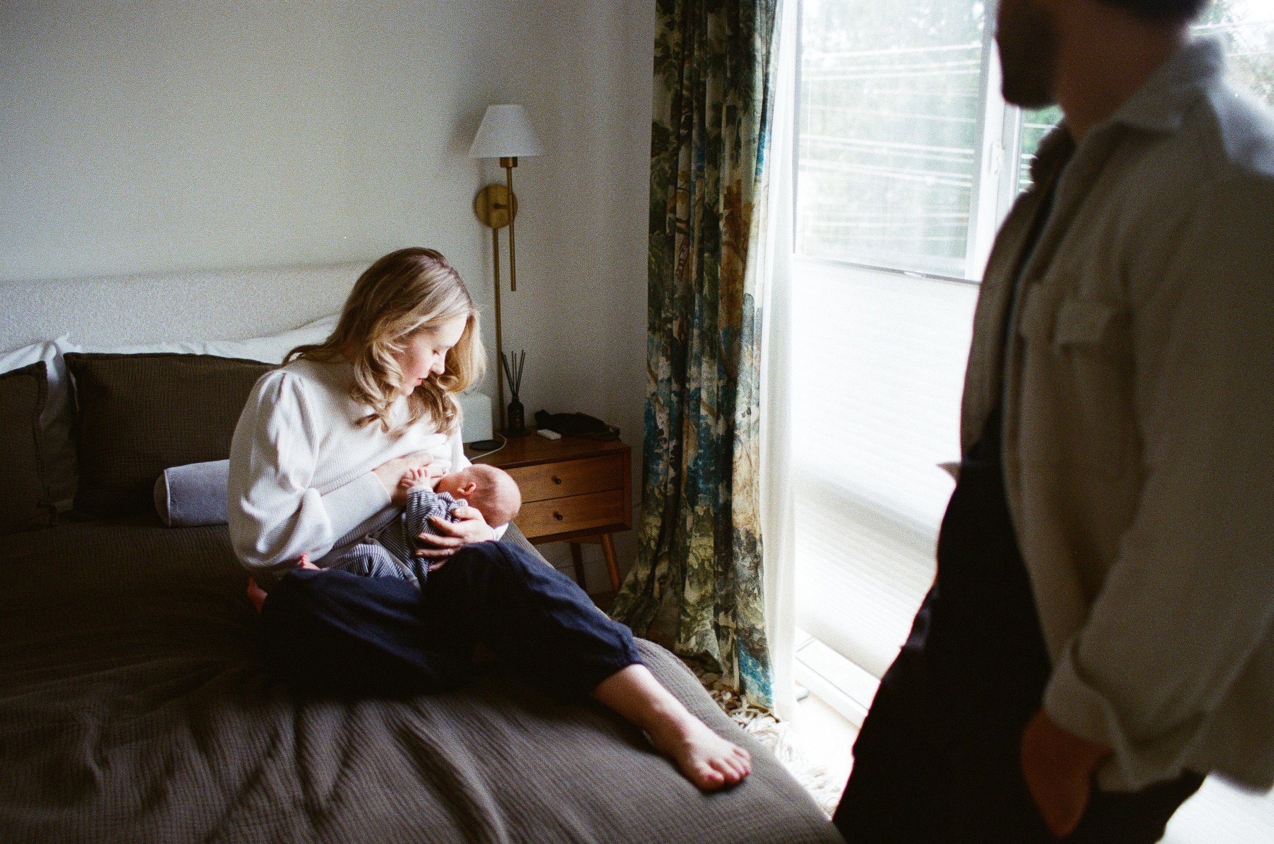 Seattle mom to a newborn baby boy breastfeeds on the bed as dad watches her lovingly.