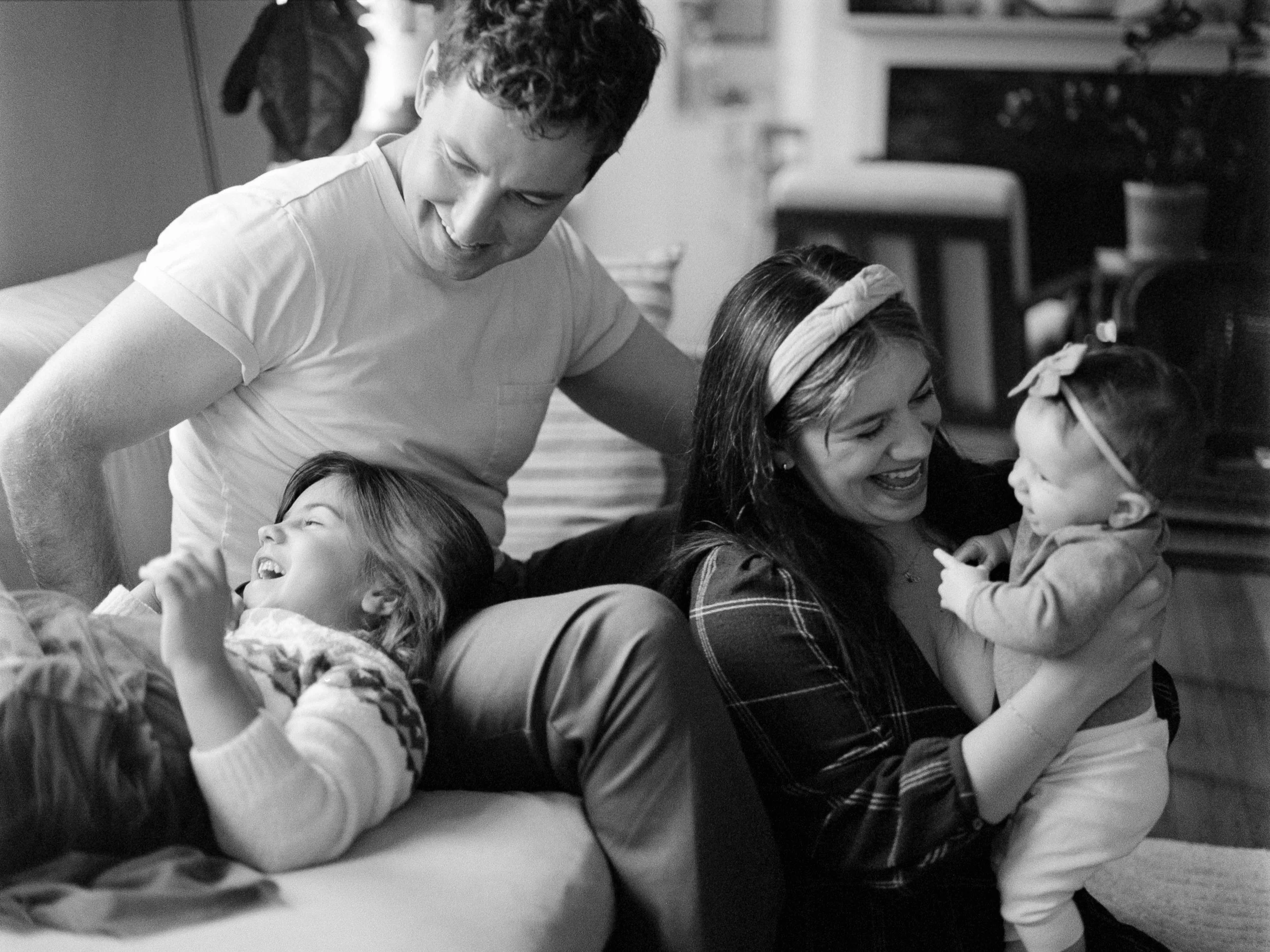 A candid, grain-textured lifestyle photograph of a family laughing on a couch. The father sits behind an older daughter lying across his lap, while the mother holds a smiling baby wearing a pink bow. Natural, documentary-style in-home portraiture.