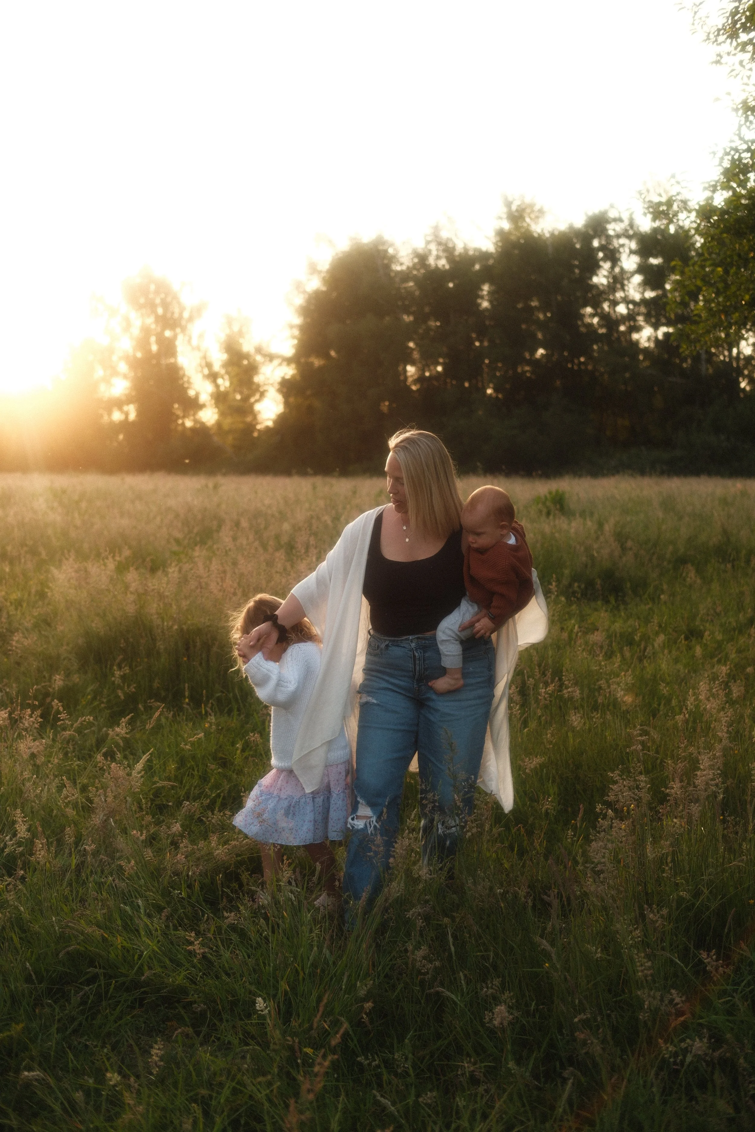 Seattle family photo of a mother standing in a field at sunset, holding her baby while her daughter smiles at the camera.