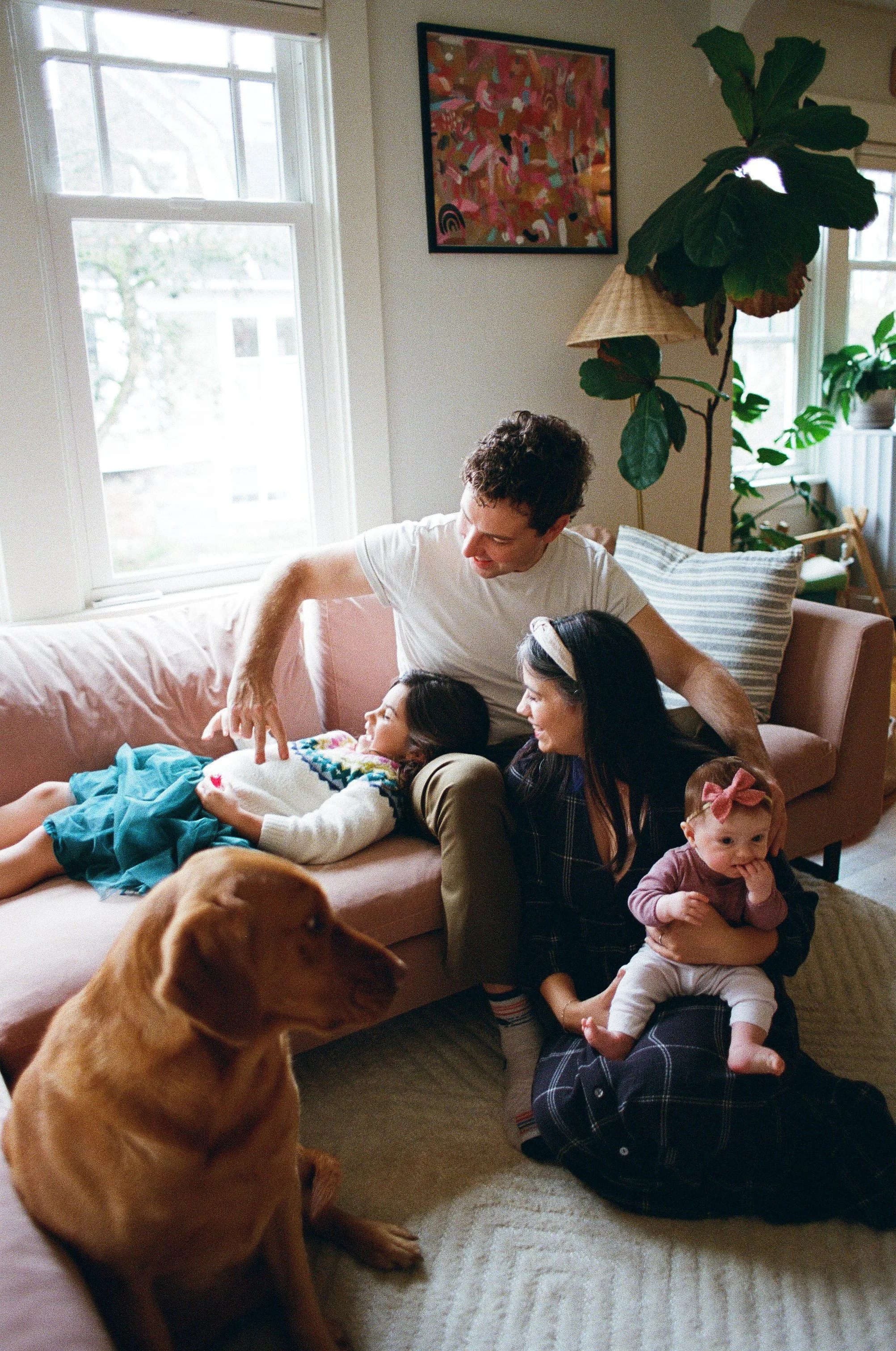 An in-home lifestyle family photography session in Seattle featuring a couple on a dusty pink sofa playing with their young daughter, while a brown dog sits on a rug in the foreground. Natural window light illuminates a cozy living room with plants.