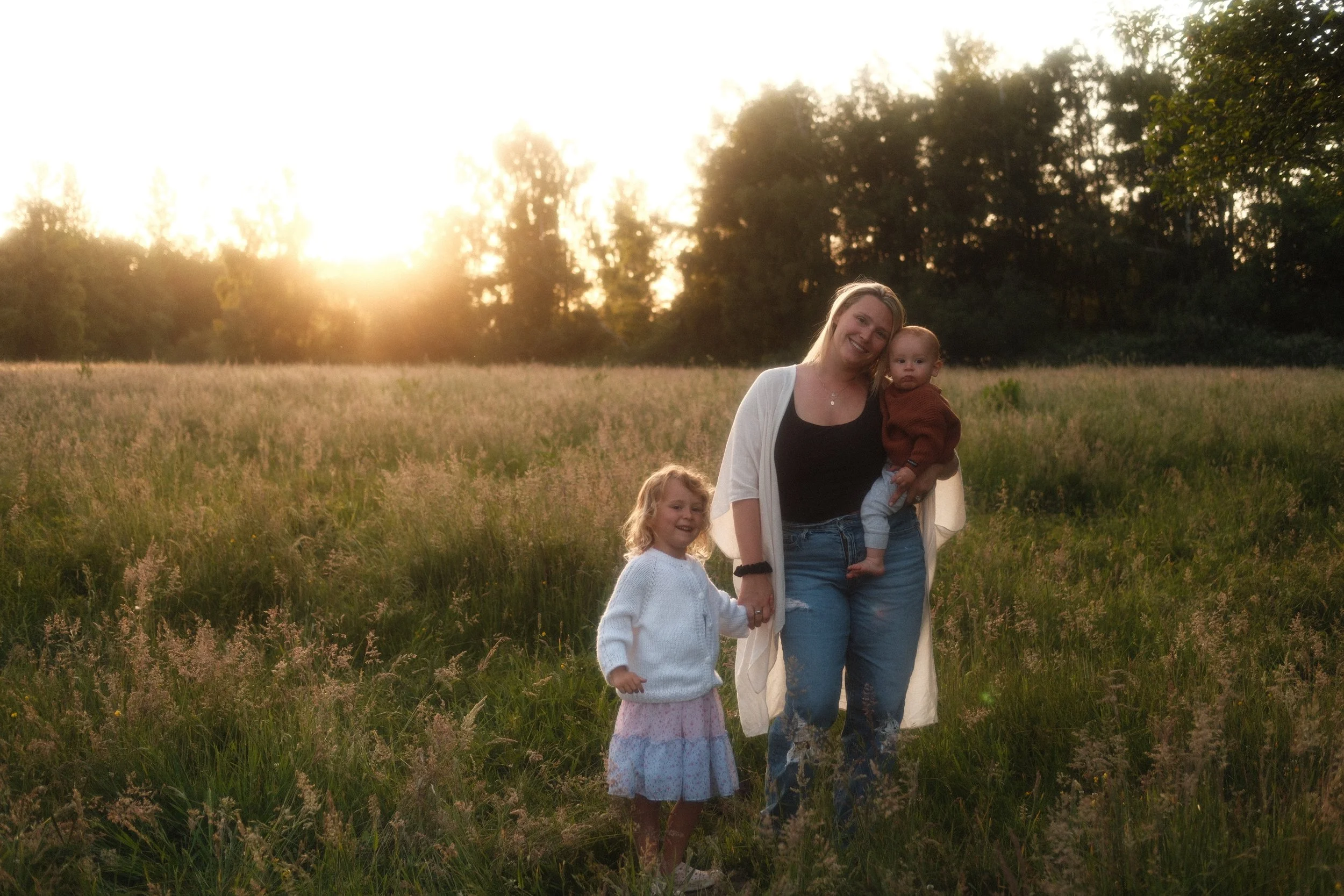 Mother holding her baby and walking hand in hand with her daughter in a sunlit Seattle field, lifestyle family photo session.