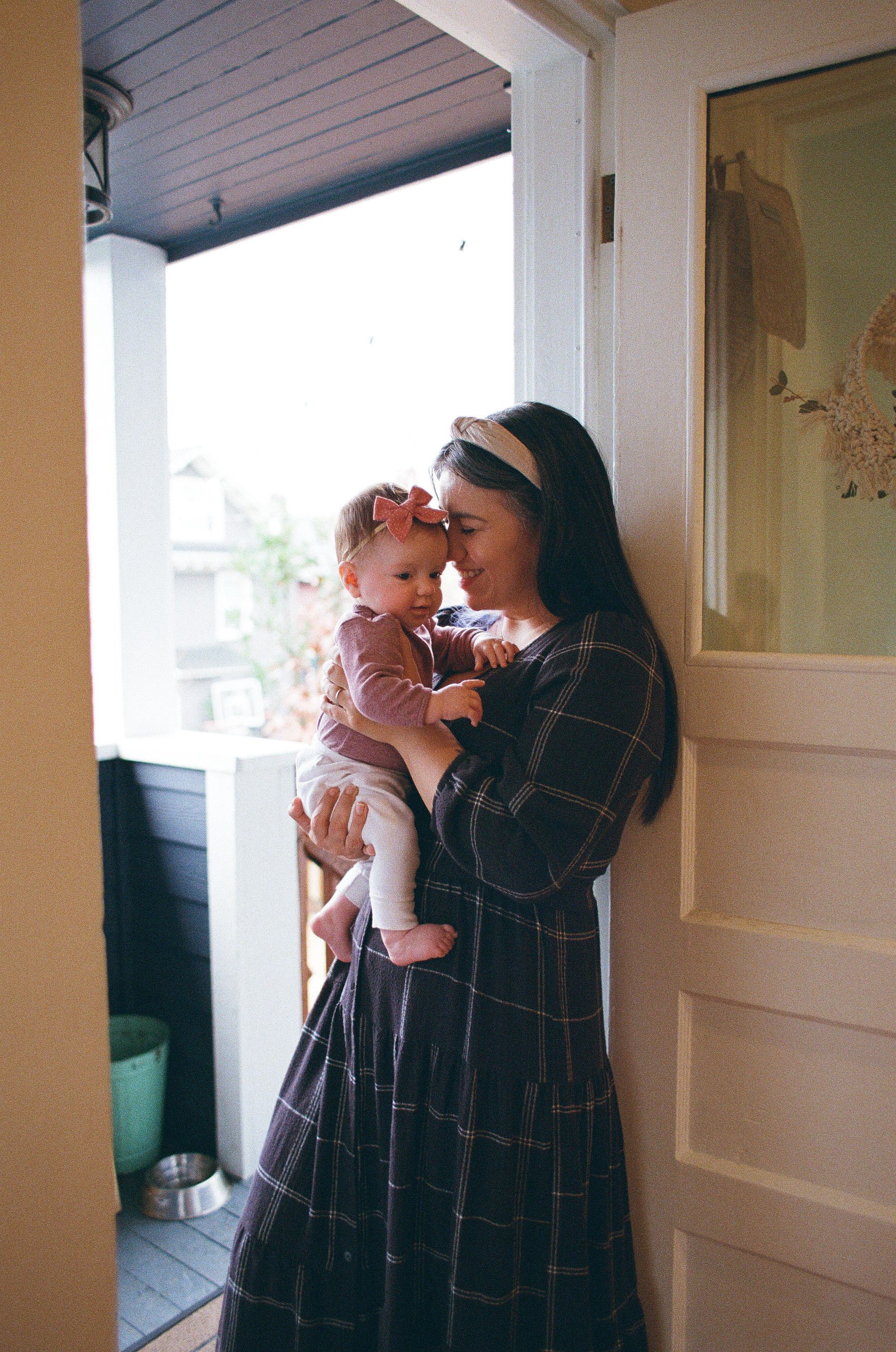 A mother in a patterned tiered dress happily holds and smiles at her baby daughter wearing a pink bow, standing by an open white front door. Lifestyle motherhood portrait photography shot in a relaxed, home setting.