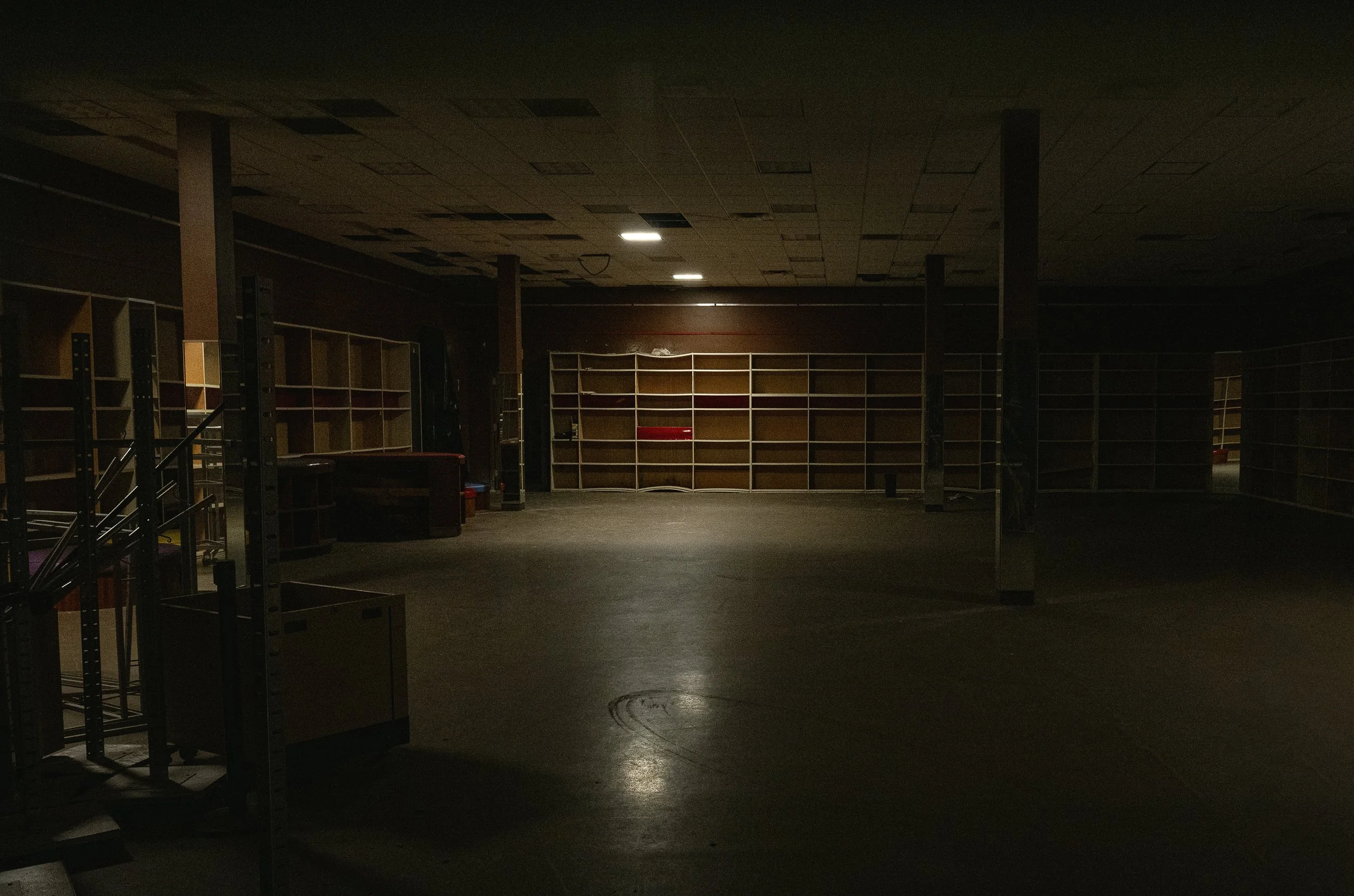 Dimly lit empty retail store or warehouse with shelves and display units, some lighting fixtures on the ceiling, and a dark floor.
