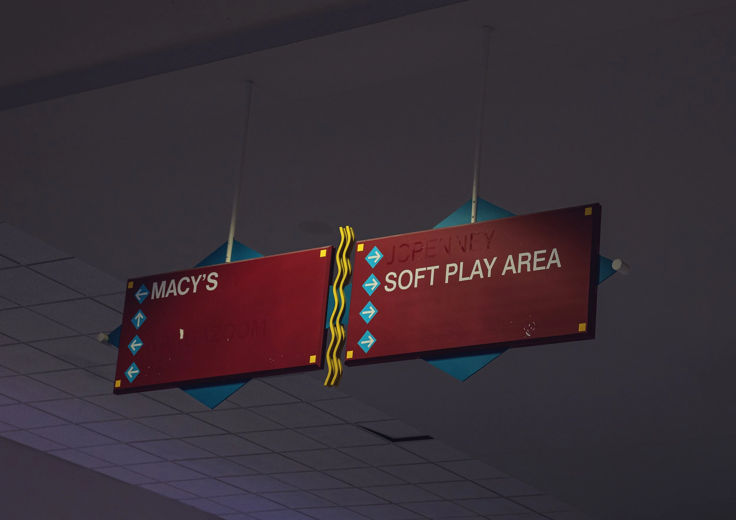 Red directional sign overhead with blue arrows pointing left and right, labeled 'Macy's,' 'Post Play Area,' and 'Soft Play Area' in an indoor setting with a tiled ceiling.