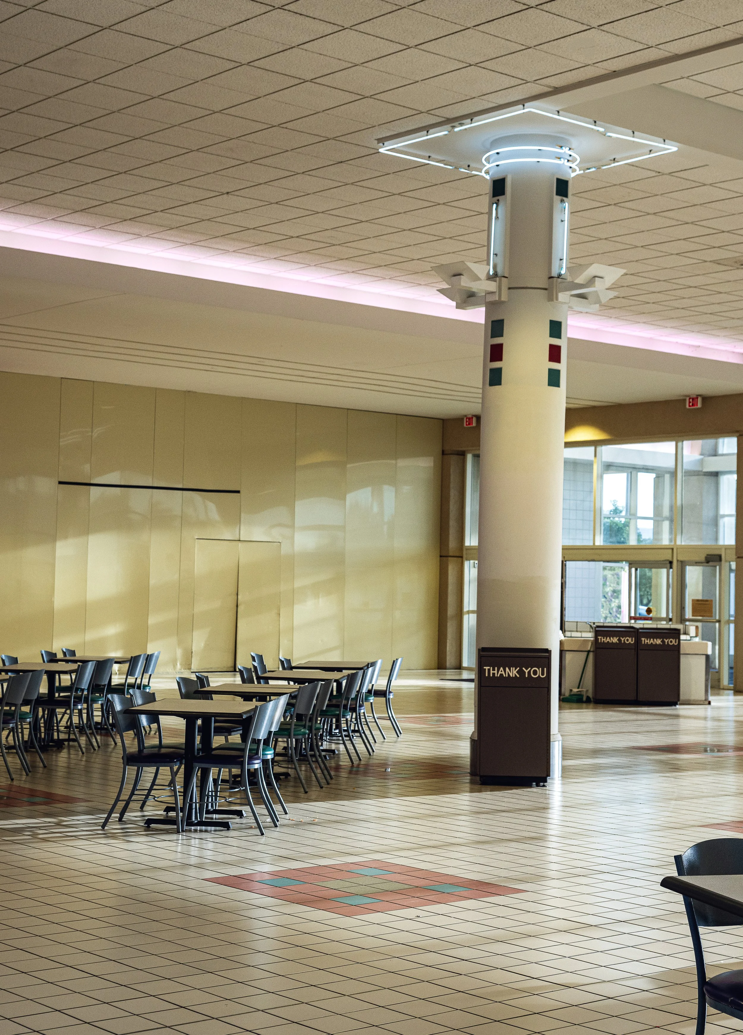 Empty indoor space with rows of tables and chairs, a large column with rectangular lights and decorations, and donation stations with 'Thank You' signs.