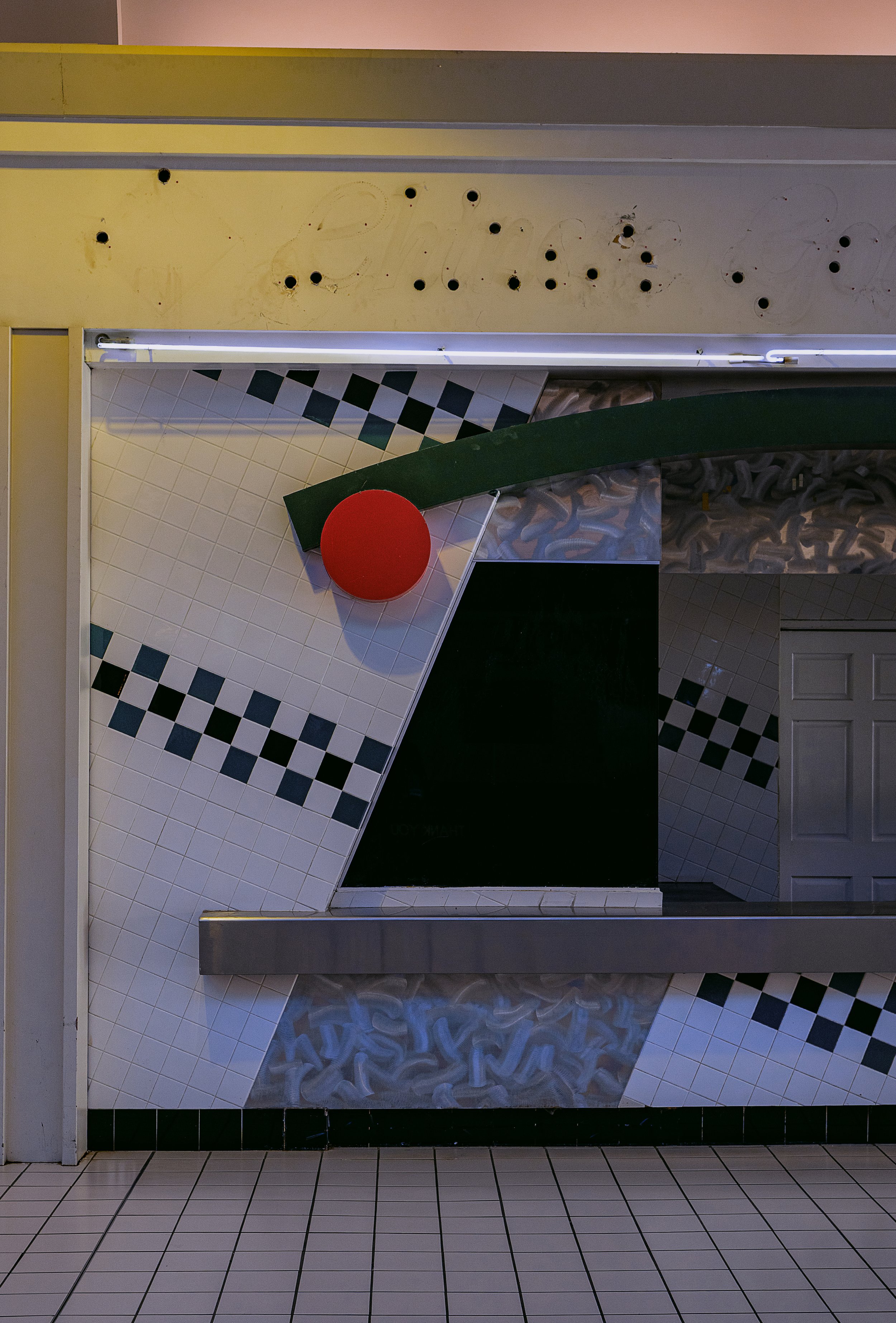 Inside a fast food restaurant booth with a worn yellow wall that has black holes and faded writing, a patterned white and black tiled wall with a red circle decoration, and a blackboard surface below. The floor is covered with white tiles with black 