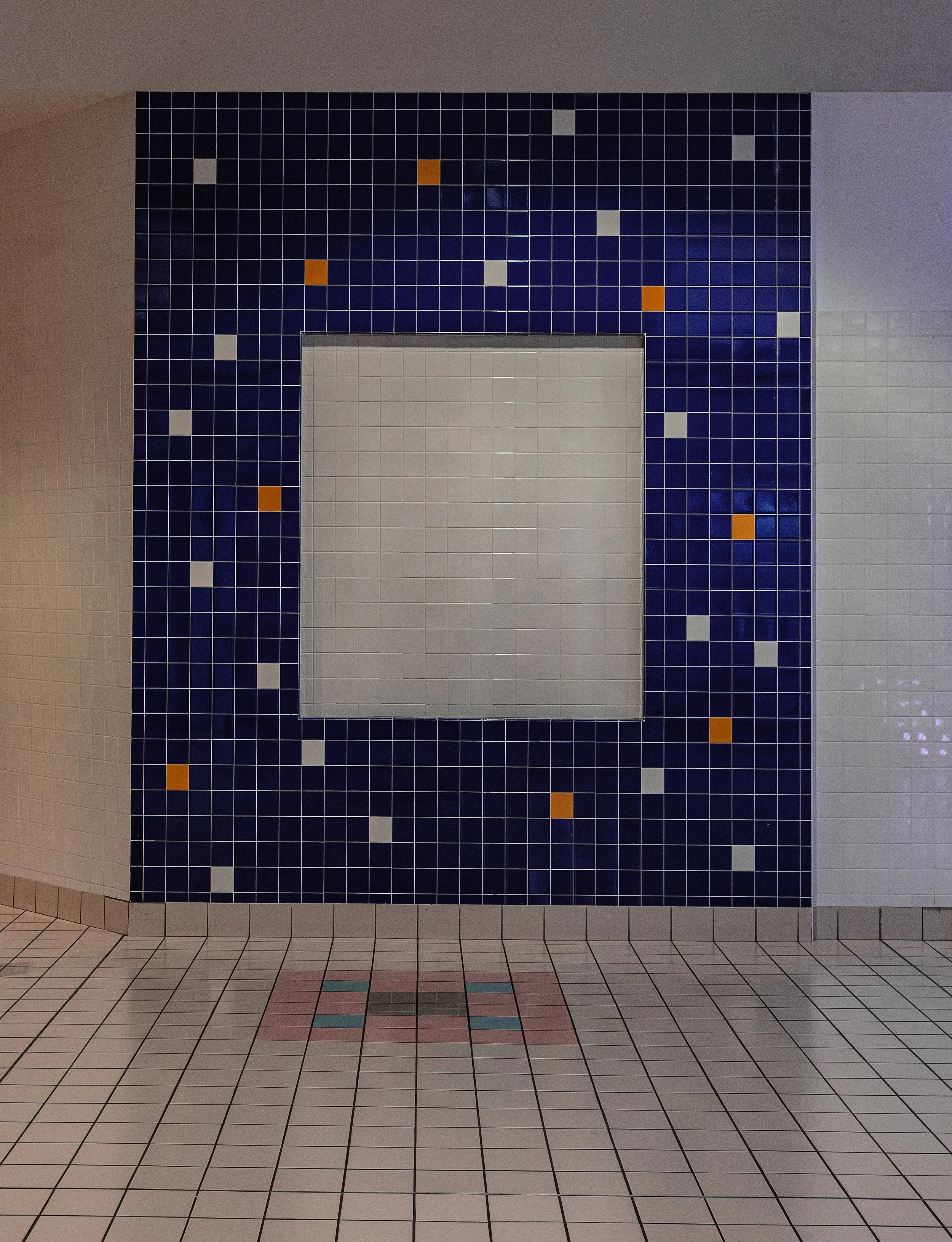A tiled wall with a geometric pattern of white, black, blue, and orange tiles in a public restroom. The lower part of the image shows a tiled floor with a similar pattern.